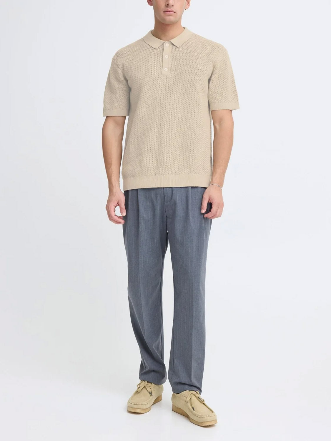 A man stands against a white background wearing the Casual Friday Cfisaksson Polo Knit in beige, styled with gray pleated trousers and light beige shoes—a smart choice for a laid-back office vibe.