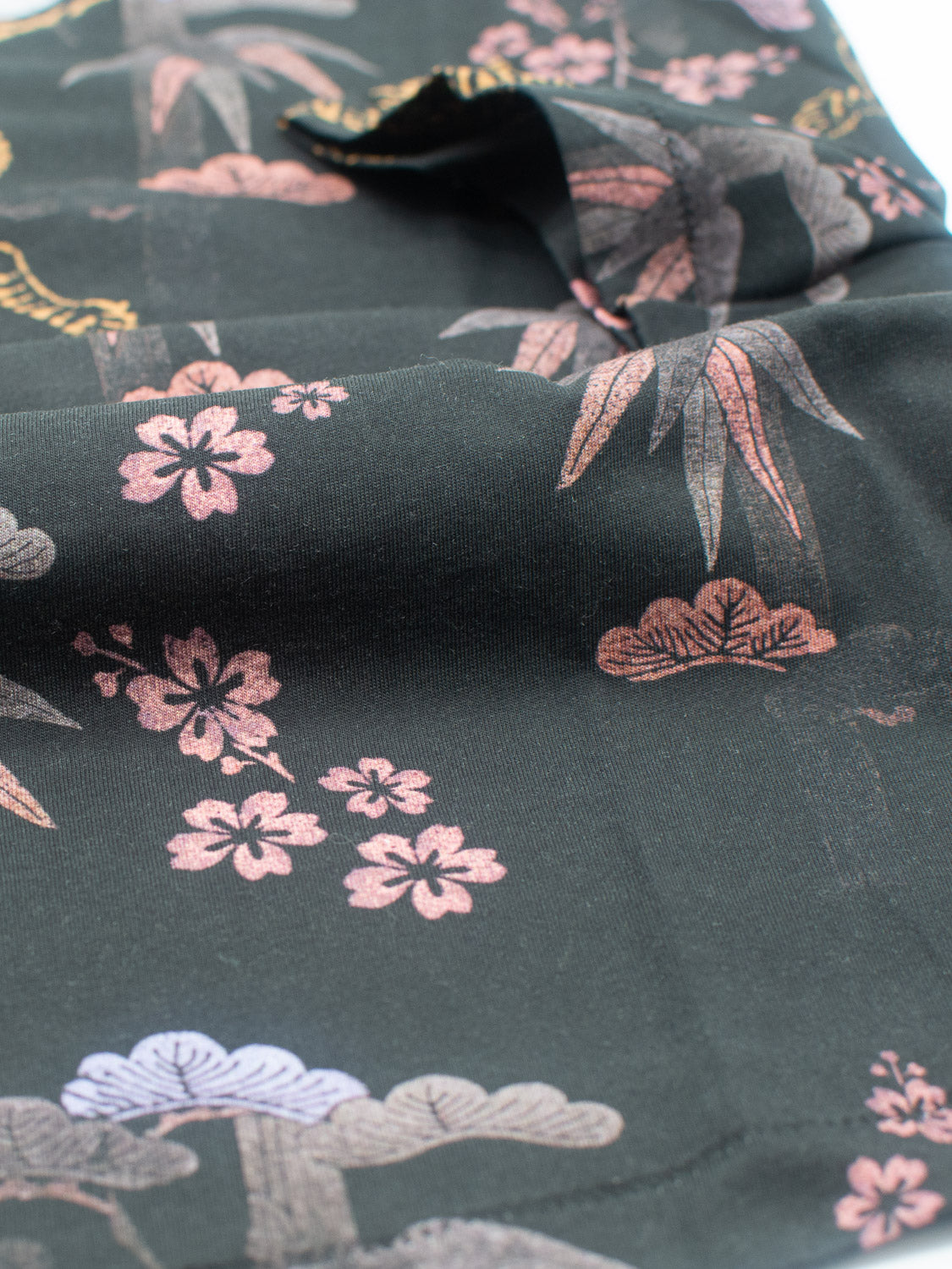 Close-up of Bali Vintage Japanese Print Tee by ID Menswear, crafted from organic cotton fabric with a black base and floral and leaf design in pink, light blue, and muted gold. The partially folded fabric creates soft curves and shadows reminiscent of vintage Japanese charm.