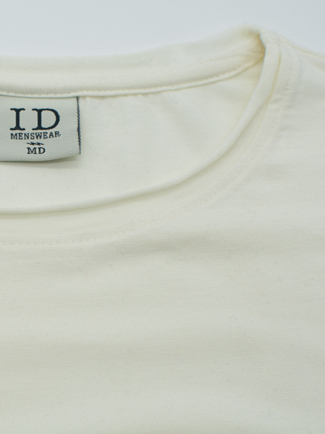 Close-up of a Bello Bamboo Blend Rolled Neck Tee by ID Menswear. The soft, textured organic cotton t-shirt has a label at the neckline reading ID MENSWEAR MD in black text and is laid flat on a white surface, embodying sustainable clothing practices.