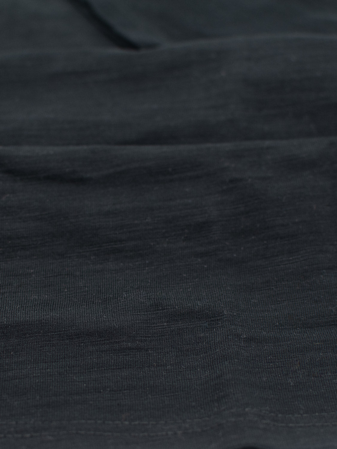 Close-up of ID Menswears Big Flip Slub Yarn Cotton Tee features a dark, textured fabric with subtle folds. Made from 100% cotton, it appears smooth and slightly shiny, reflecting minimal light and creating gentle shading variations reminiscent of a slub yarn t-shirt.