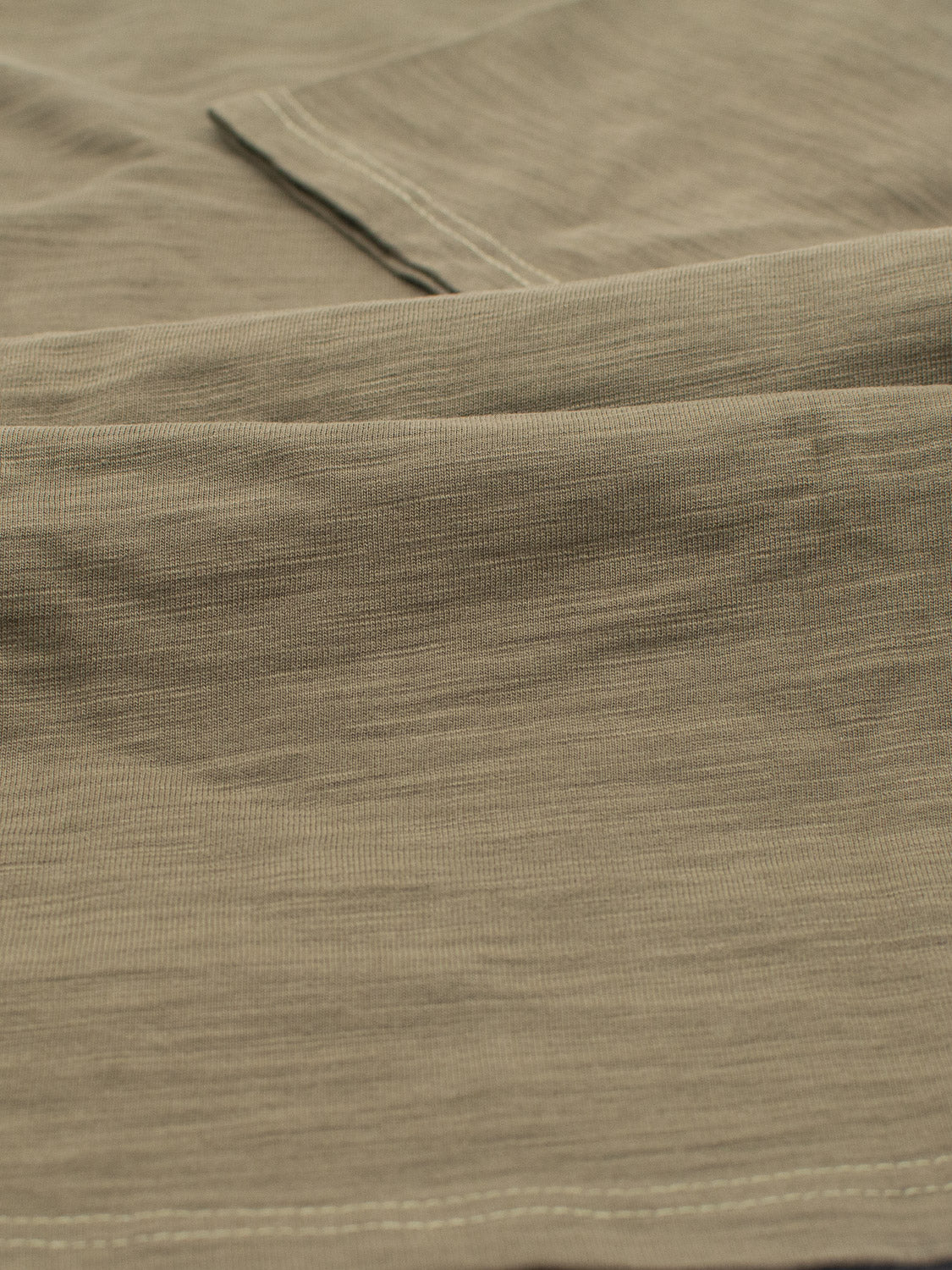 Close-up of the Big Flip Slub Yarn Cotton Tee by ID Menswear shows light brown 100% cotton fabric with a visible texture and stitching. The soft, slightly wrinkled material resembles a slub yarn T-shirt, featuring a folded edge running across the image.