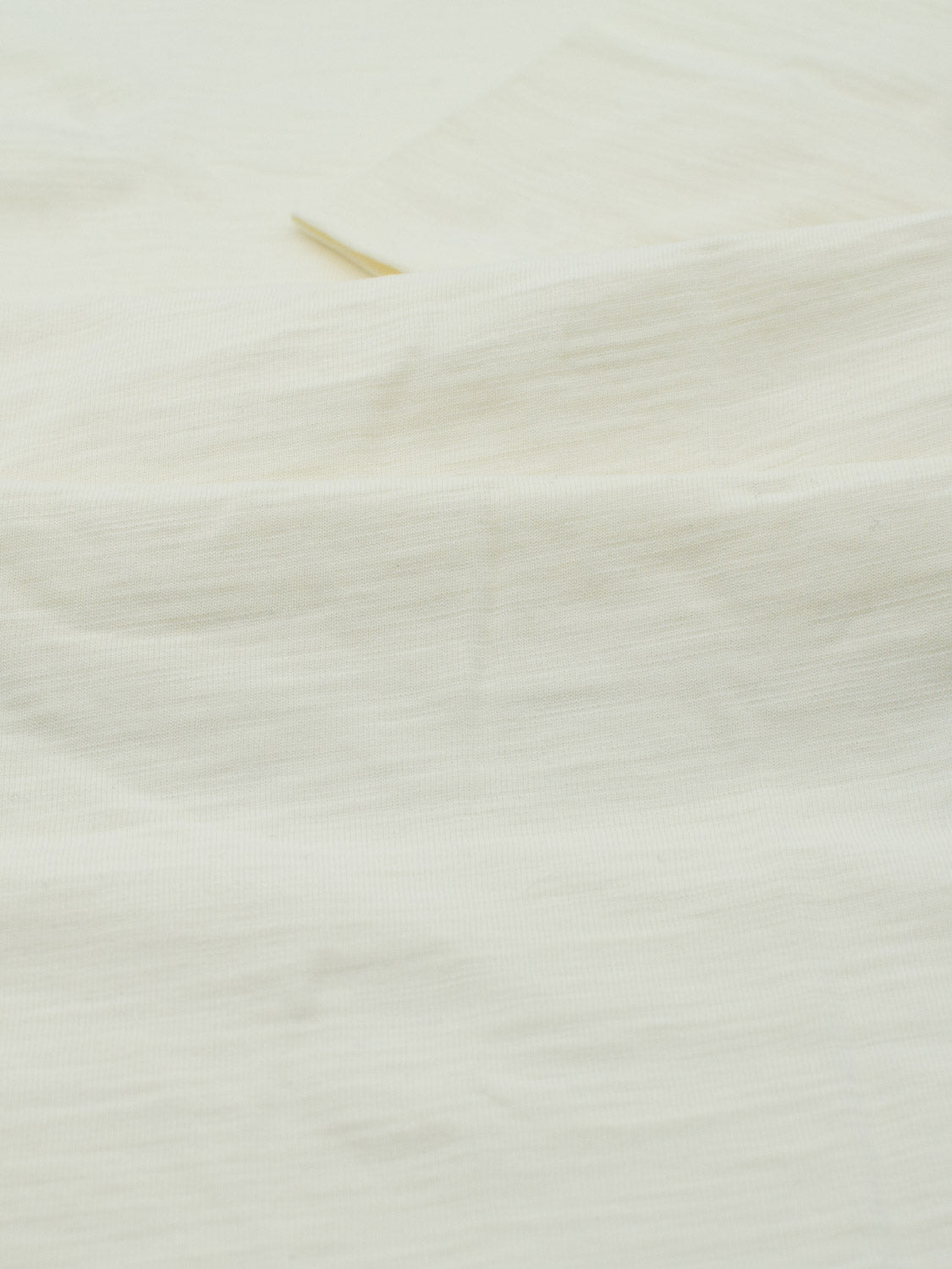 Close-up of a plain, light beige Big Flip Slub Yarn Cotton Tee by ID Menswear. Crafted from 100% cotton, the fabric is slightly textured with a soft drape, showing faint lines and creases. Minimal shadowing suggests overhead lighting.