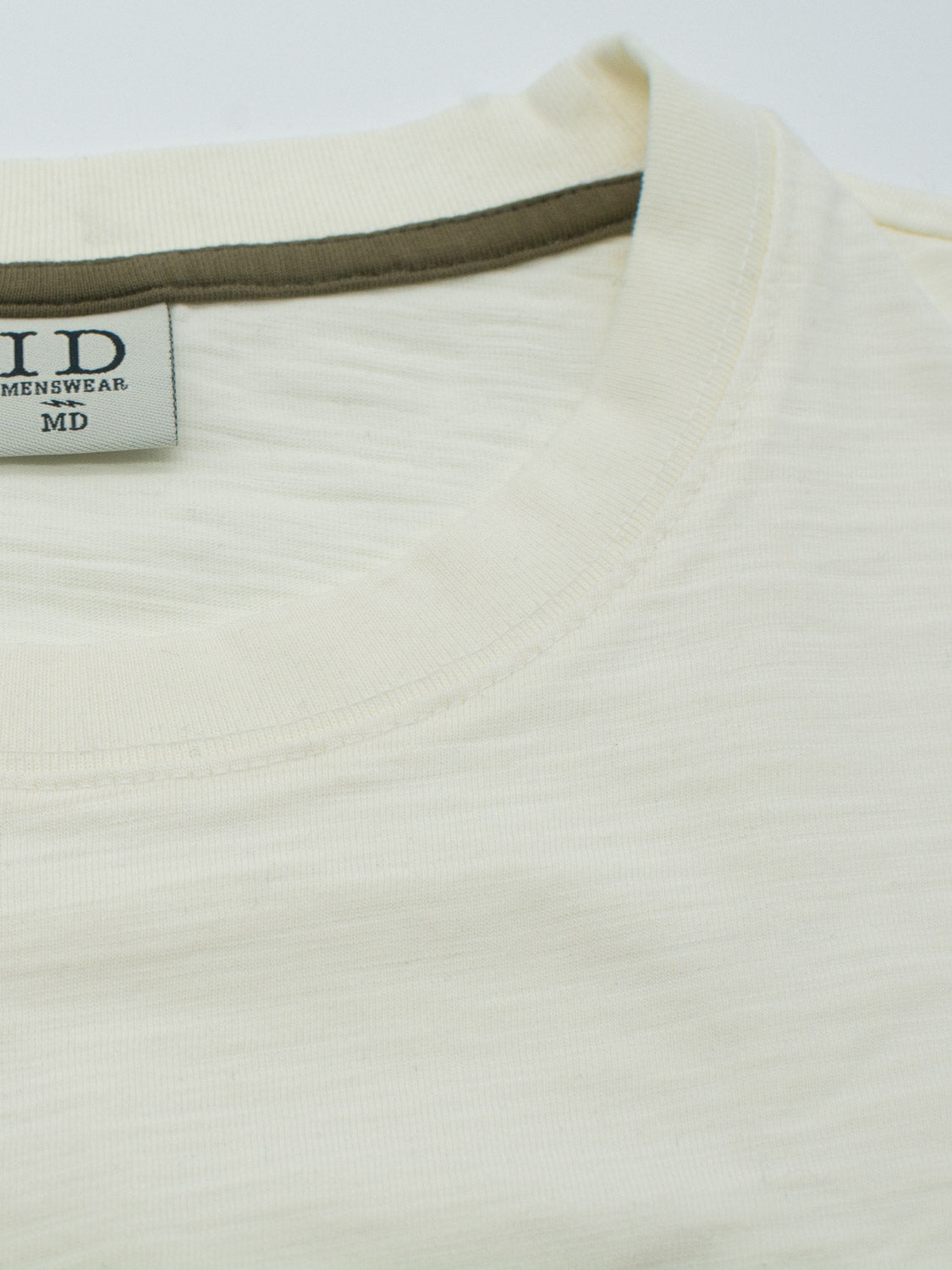 A close-up of the cream-colored Big Flip Slub Yarn Cotton Tee from ID Menswear shows the MD size label. It features a simple round neckline with a darker trim and a boxy fit. The shirt, made from 100% cotton, has a soft, lightweight texture.