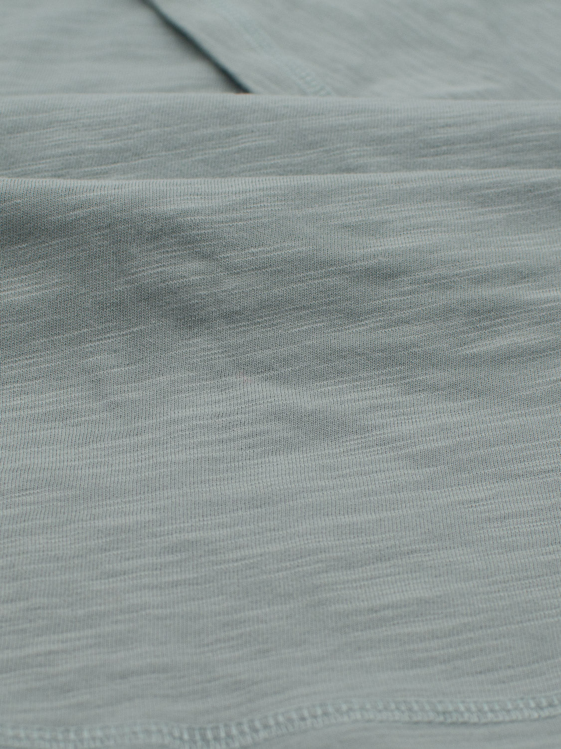 Close-up of light teal slub cotton fabric from ID Menswears Da Nang Rolled Neck Slub Tee, featuring a soft, smooth texture with subtle horizontal lines. The lightweight material is slightly crinkled with fine stitching at the bottom edge—ideal for casual wear.