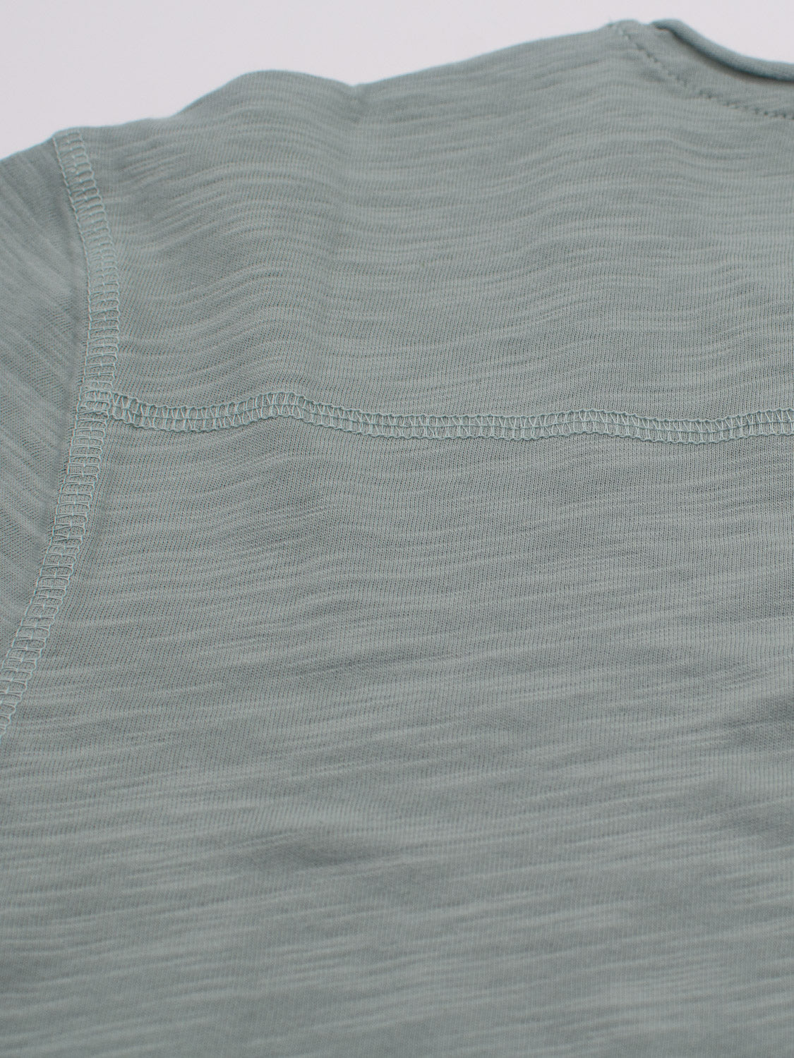 Close-up of the back of a Da Nang Rolled Neck Slub Tee by ID Menswear in light gray-green, highlighting detailed stitching across the upper back and shoulders. The slub cotton has a subtle texture, ideal for casual elegance enthusiasts.