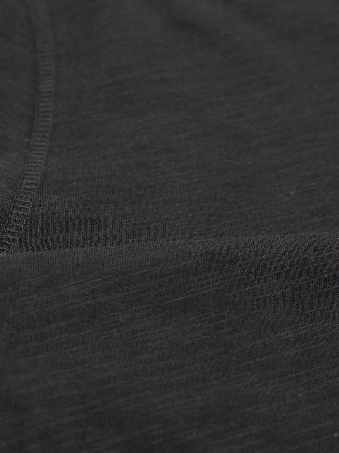 Close-up of black slub cotton fabric from the Da Nang Rolled Neck Slub Tee by ID Menswear. The material is soft and slightly heathered, ideal for casual wear, with visible stitching and texture, plus a seam running across the image.