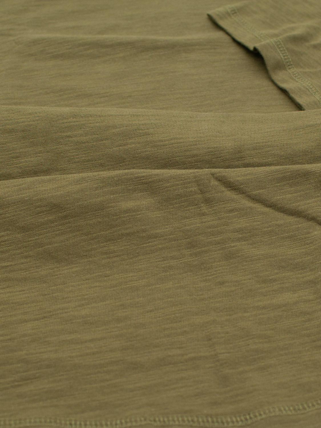 A close-up shows a textured olive green fabric with visible seams and subtle wrinkles, similar to slub cotton, likely from ID Menswears Da Nang Rolled Neck Slub Tee, lying flat.