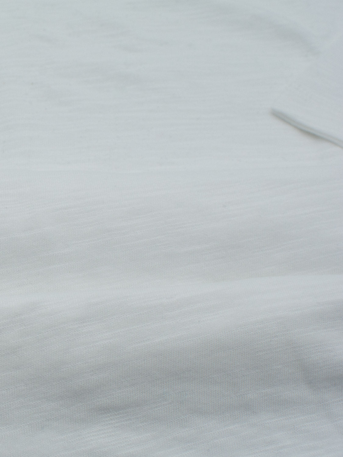 Close-up of light gray fabric with a smooth, slightly textured surface. The top right corner shows a folded edge, suggesting it might be slub cotton from ID Menswears Da Nang Rolled Neck Slub Tee.