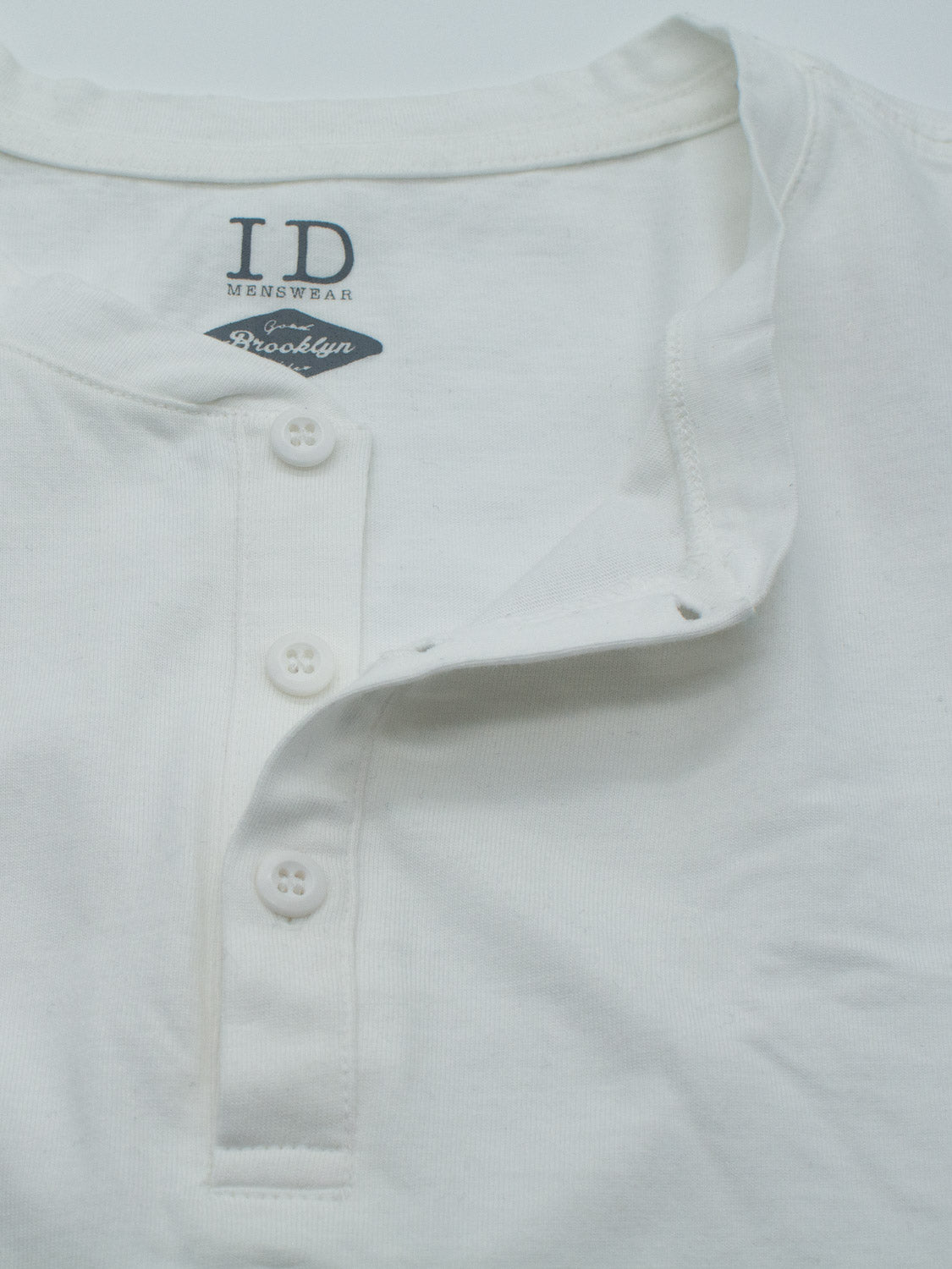 Close-up of the Champion Organic Cotton Henley by ID Menswear, showcasing a classic silhouette with three buttons and a fold-over collar. The label reads ID Menswear Brooklyn in dark print. Made from organic cotton jersey, the fabric is soft with a slight texture.
