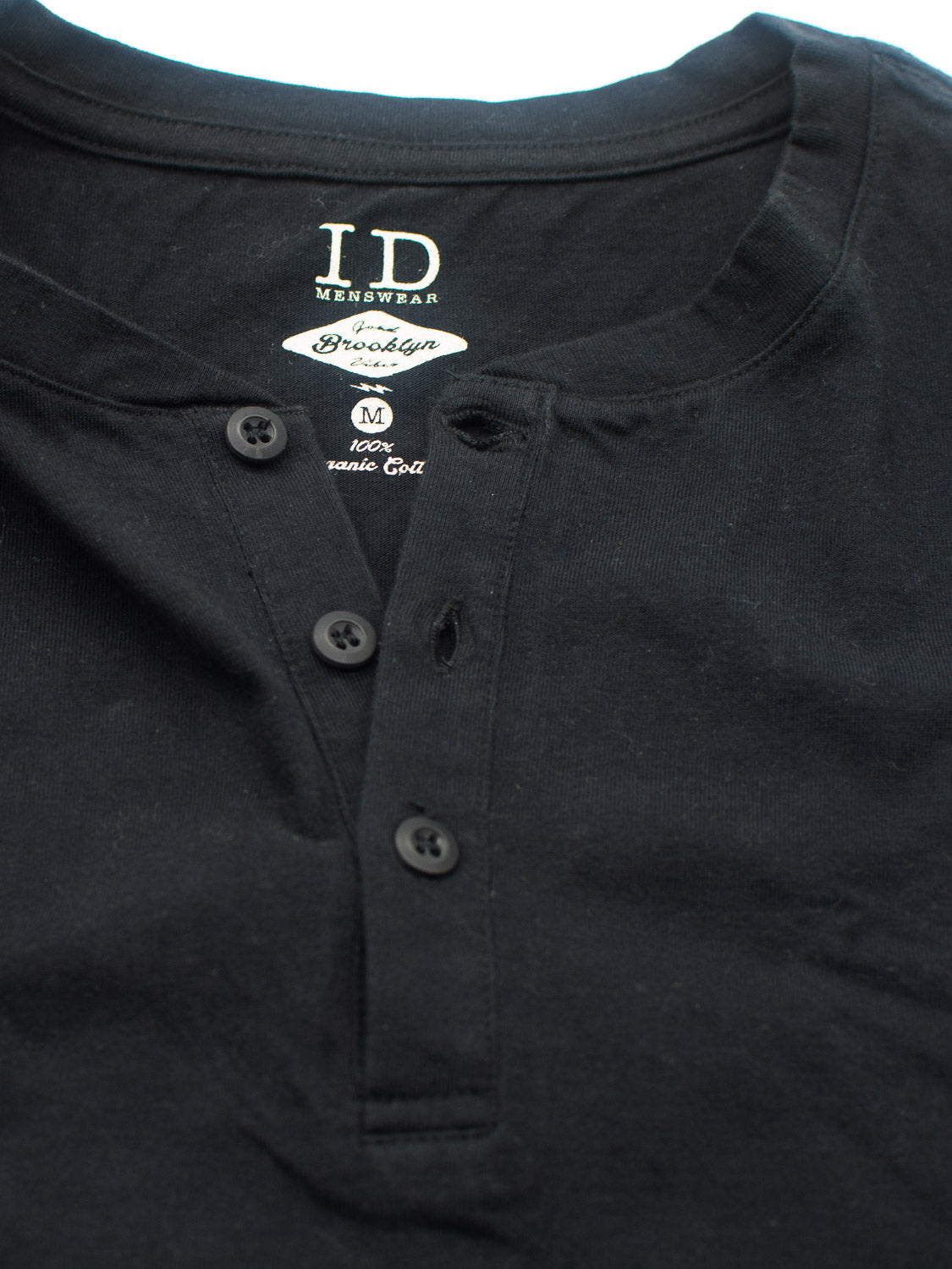 A close-up of the Champion Organic Cotton Henley by ID Menswear in black shows its classic men’s silhouette with three visible buttons. The inner tag indicates its a medium size, crafted from 100% organic cotton jersey.