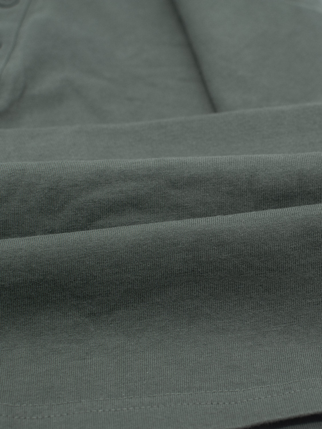 Close-up of folded dark green Champion Organic Cotton Henley by ID Menswear, showcasing a texture similar to organic cotton jersey. The material appears soft and thick, with neatly creased folds that accentuate its quality and classic silhouette.