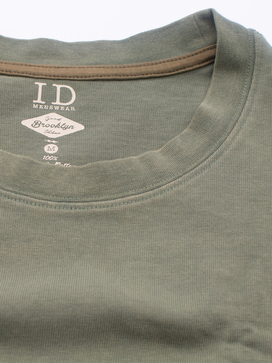 A close-up shows a green Freeman Organic Vintage Wash Tee by ID Menswear, size M. Brooklyn Grade is inside the collar. Made from soft organic cotton, the fabric has a slight ribbing for a vintage worn look.