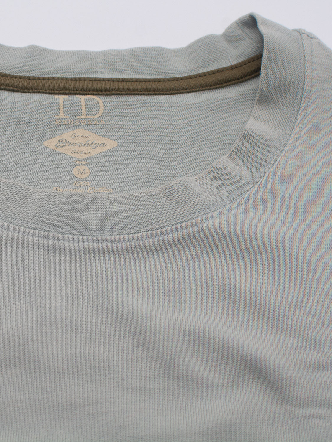 Close-up of two Freeman Organic Vintage Wash Tees by ID Menswear, with the top in light grey and the bottom in olive green. The grey shirt has a size M Brooklyn tag. Made from soft, ribbed organic cotton, they offer a vintage worn look.