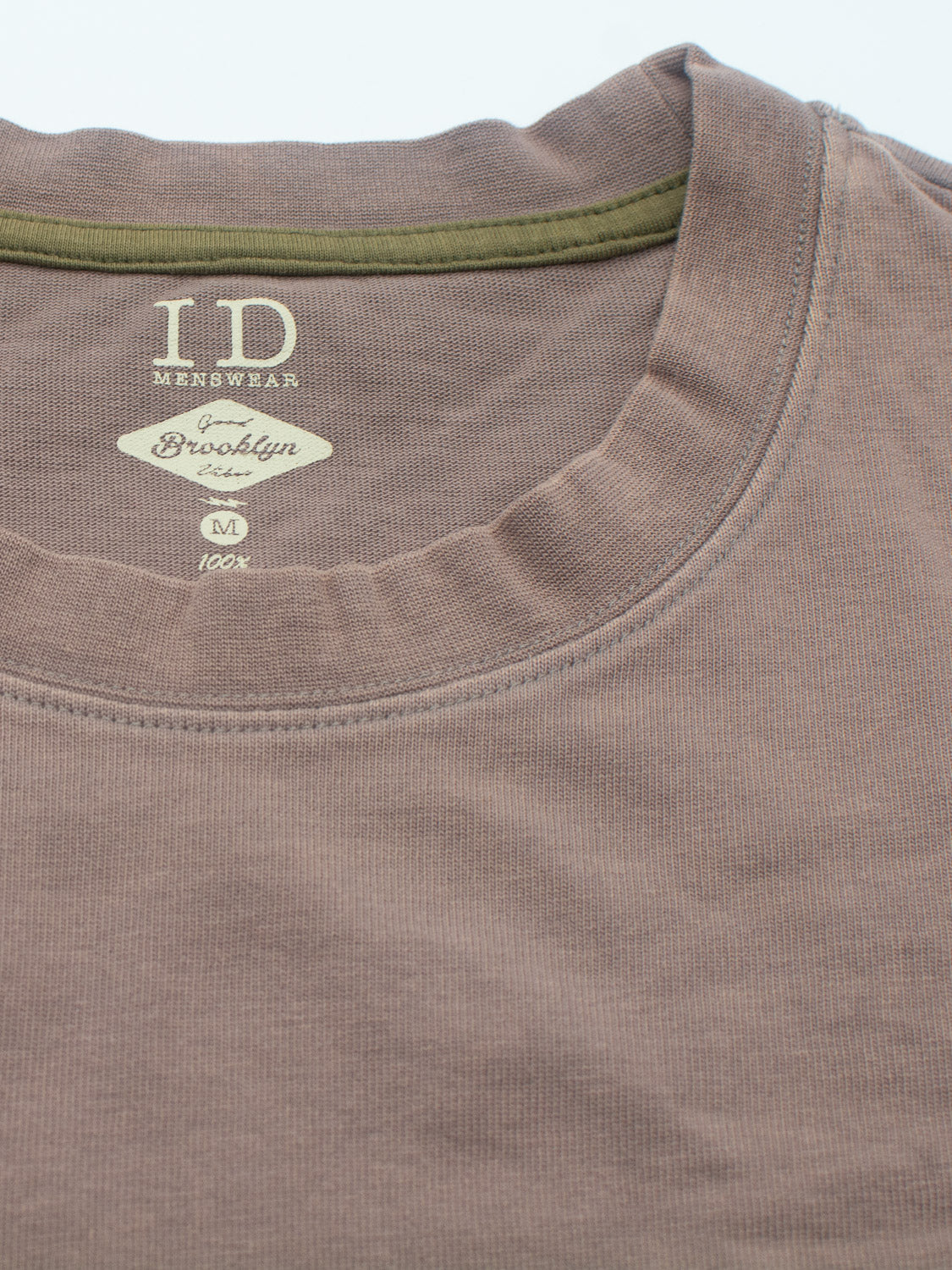 Close-up of the Freeman Organic Vintage Wash Tee by ID Menswear, a brown T-shirt featuring an olive green neckline and vintage worn look. Crafted from 100% organic cotton with a boxy fit, it includes Brooklyn text on the label and is indicated as medium size.