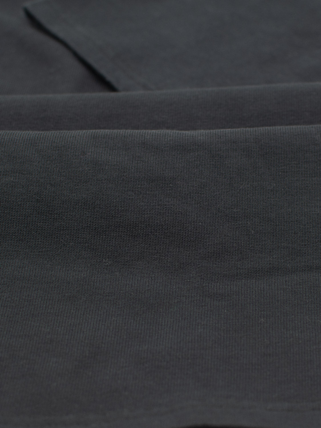 A close-up of dark gray, textured fabric evokes the City Organic Cotton Tee by ID Menswear. Crafted from organic cotton, two overlapping layers create subtle folds and shadows, with a fine weave enhancing its smooth look.
