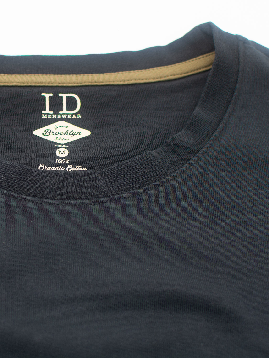 Close-up of a black City Organic Cotton Tee for men by ID Menswear features a round neckline, Brooklyn label, 100% organic cotton, slim fit design, size M, and brown inner neck fabric.