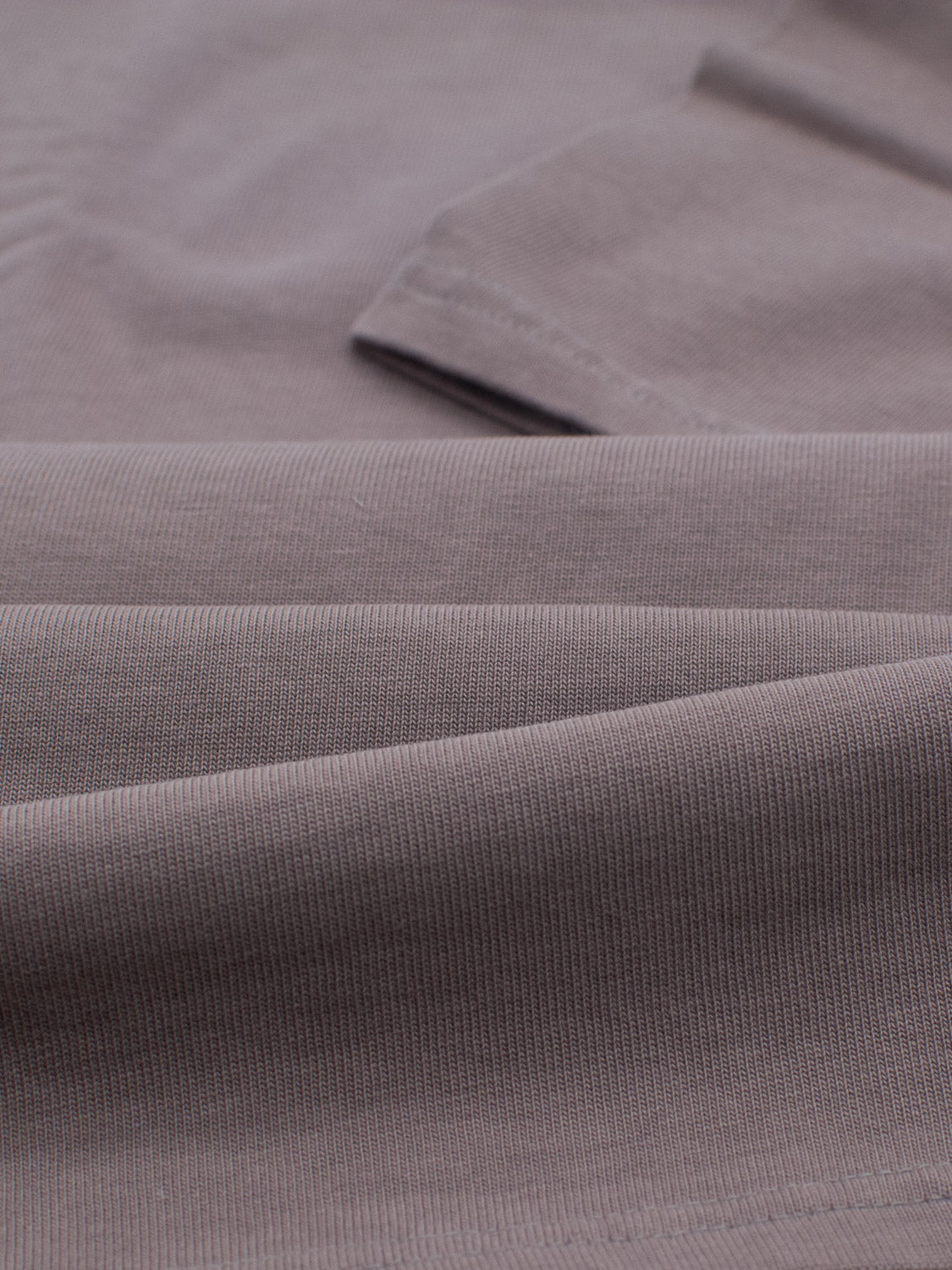 Close-up of a folded, ribbed fabric in muted taupe, crafted from organic cotton. The texture shows a subtle linear pattern and smooth finish reminiscent of the City Organic Cotton Tee by ID Menswear.