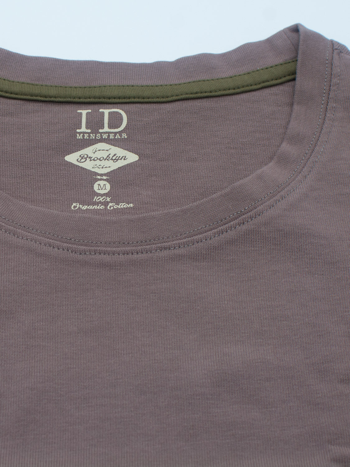 A folded brown City Organic Cotton Tee with a green collar, labeled ID Menswear, Brooklyn Co., shows slim fit, size M, and 100% organic cotton in white text.