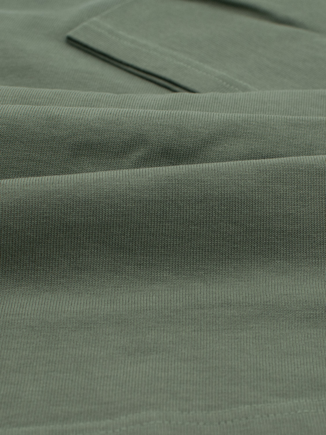 Close-up of the City Organic Cotton Tee by ID Menswear reveals a soft, textured look in green ribbed fabric with overlapping folds. Made from organic cotton, it showcases small parallel lines that add subtle pattern and depth, ideal for city wear.