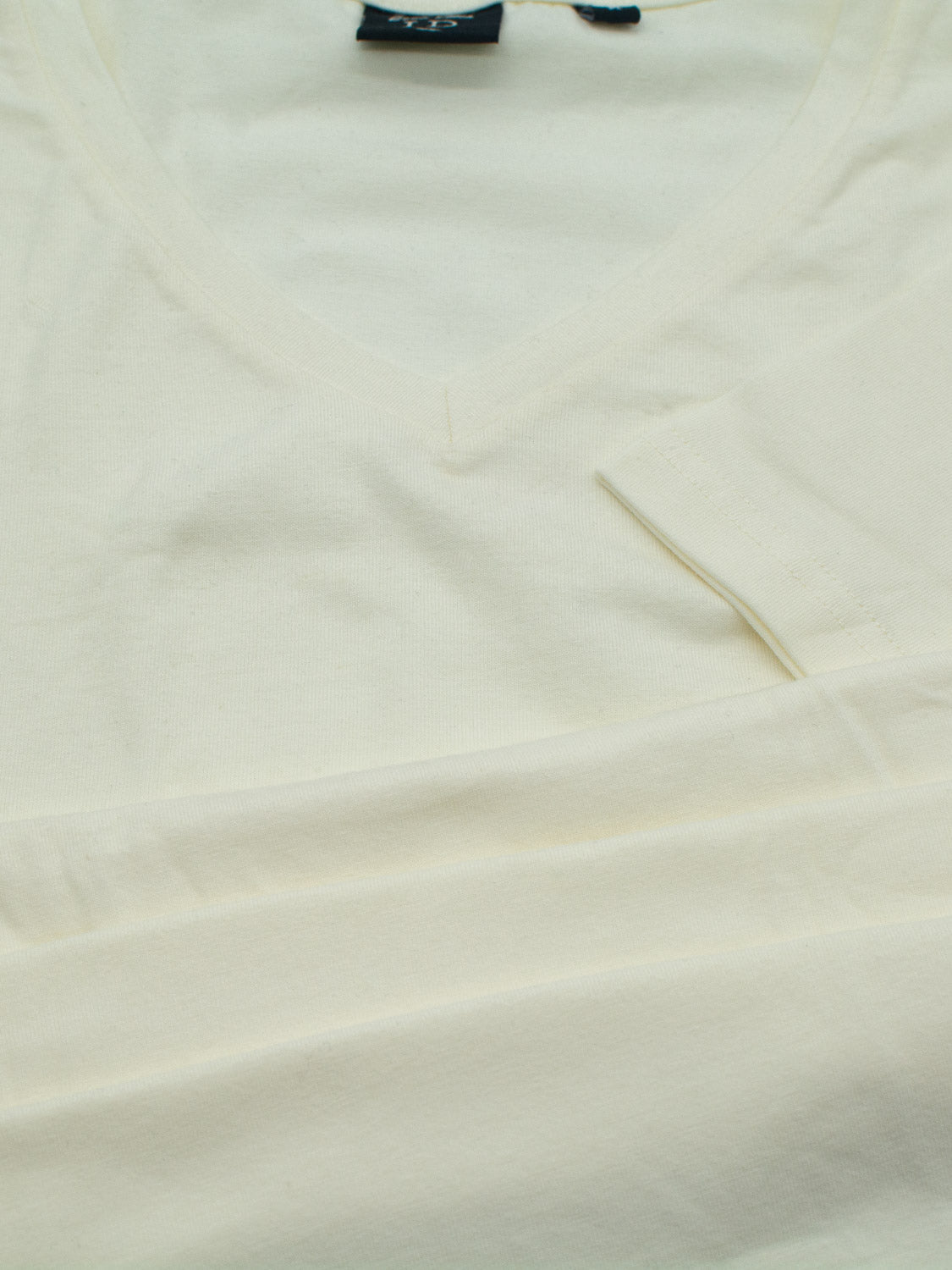 A cream Form V Neck Tee from ID Menswear is laid flat, showcasing its form-fitting style. The sleeve peeks into the foreground, with a dark clothing tag partially visible near the collar.