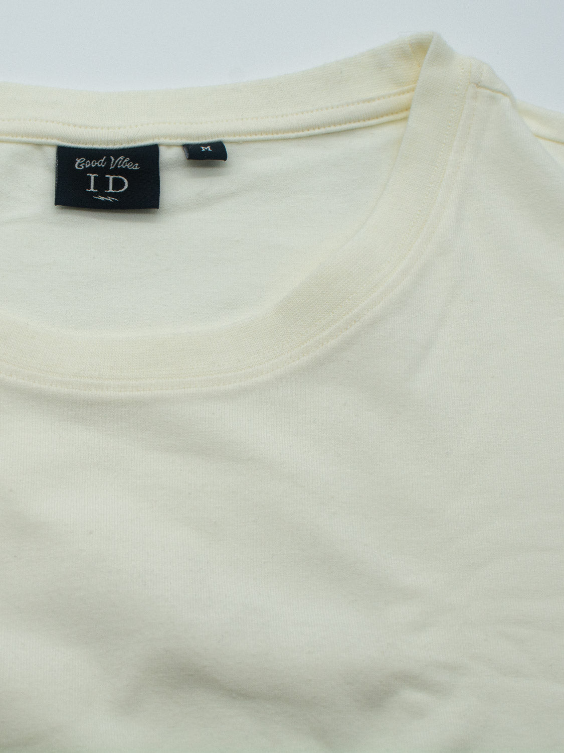 A beige Classic Crew Neck Tee from ID Menswear, crafted from soft cotton-spandex fabric, lays flat. It features a regular fit with a round neck and a visible tag on the collar adorned with text and symbols.