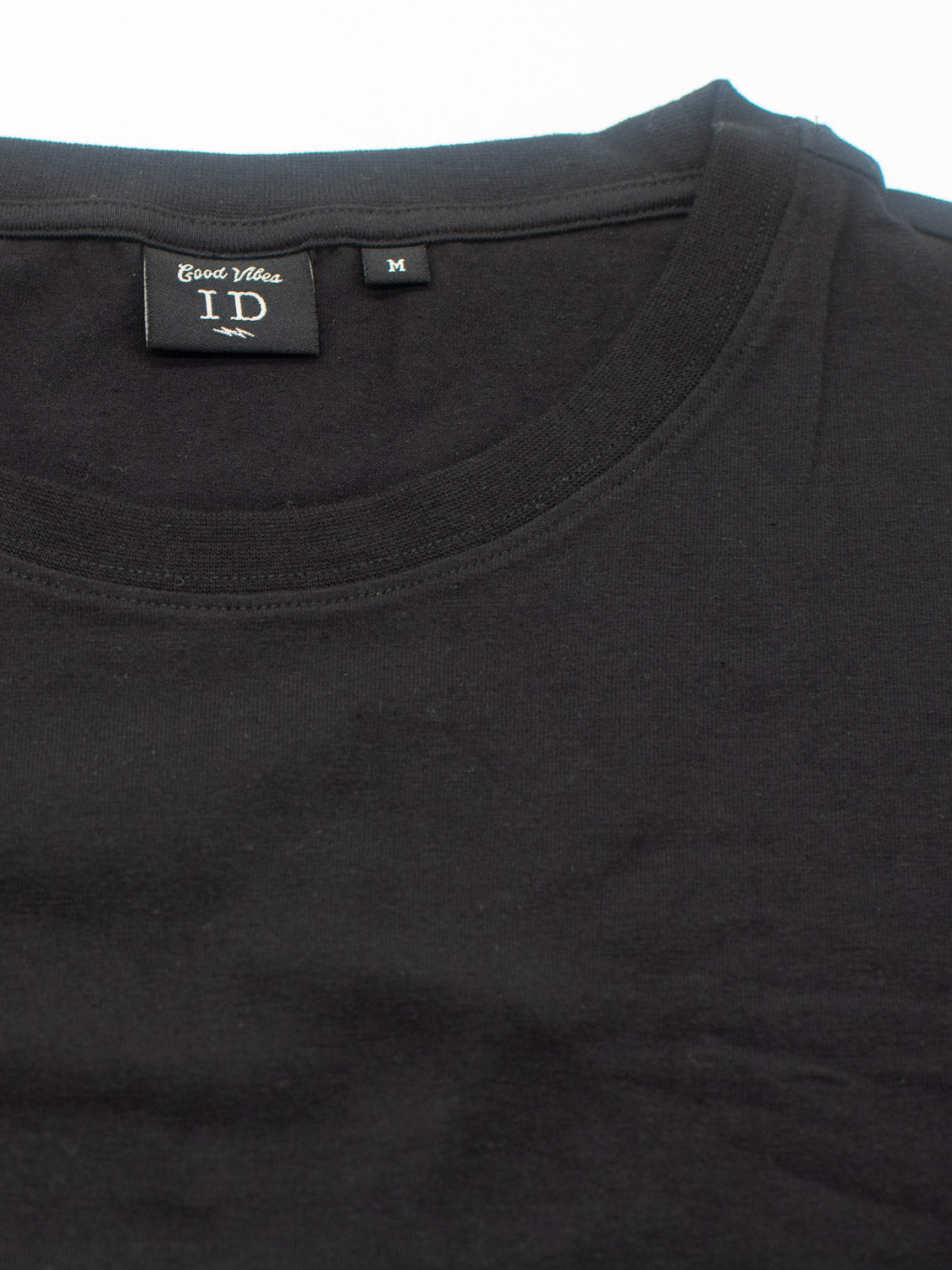 Close-up of a plain black Classic Crew Neck Tee by ID Menswear, featuring a round neck and made from cotton-spandex fabric. The inner label reads Good Vibes, indicating a Medium size for a comfortable regular fit.