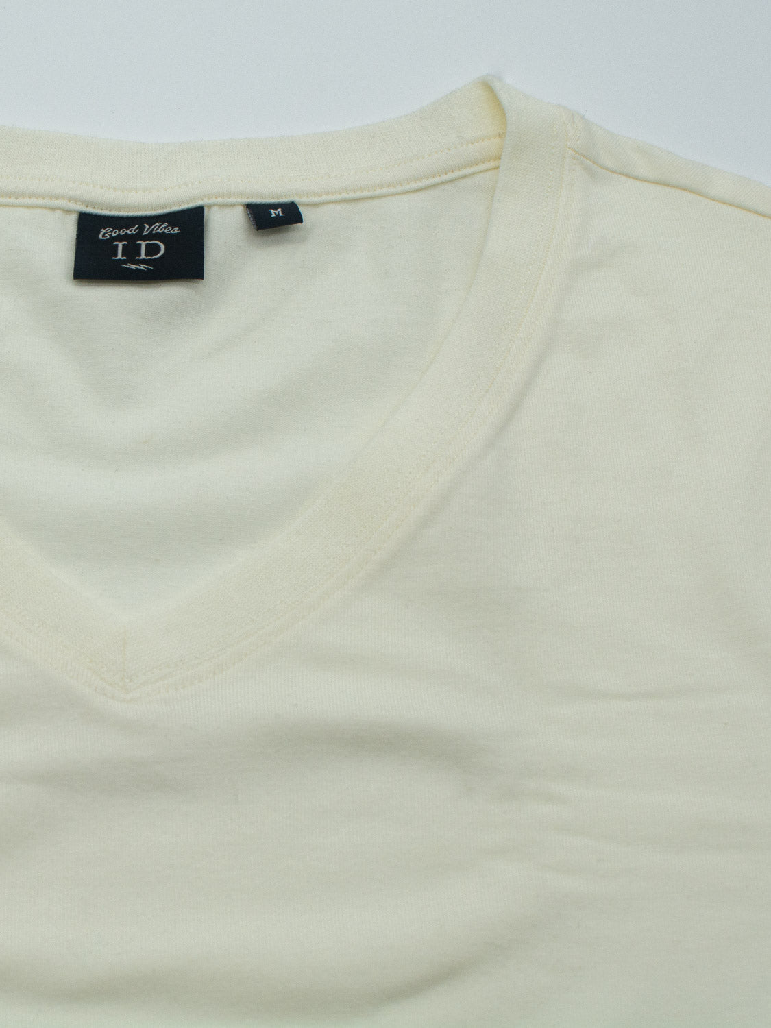 Close-up of a beige Classic V Neck Tee, a wardrobe essential with its cotton-spandex blend. A small black tag near the collar displays size M and the brand name ID Menswear. The fabric appears soft and slightly textured.