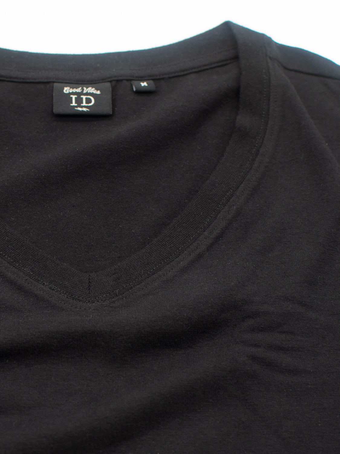 Close-up of a black V-neck t-shirt from ID Menswears Classic V Neck Tee. The cotton-spandex blend fabric is smooth and stretchy, with a plain design. Displayed flat, you can see the neckline and shoulder seam clearly. The inside label reads ID and Cool Vibe.