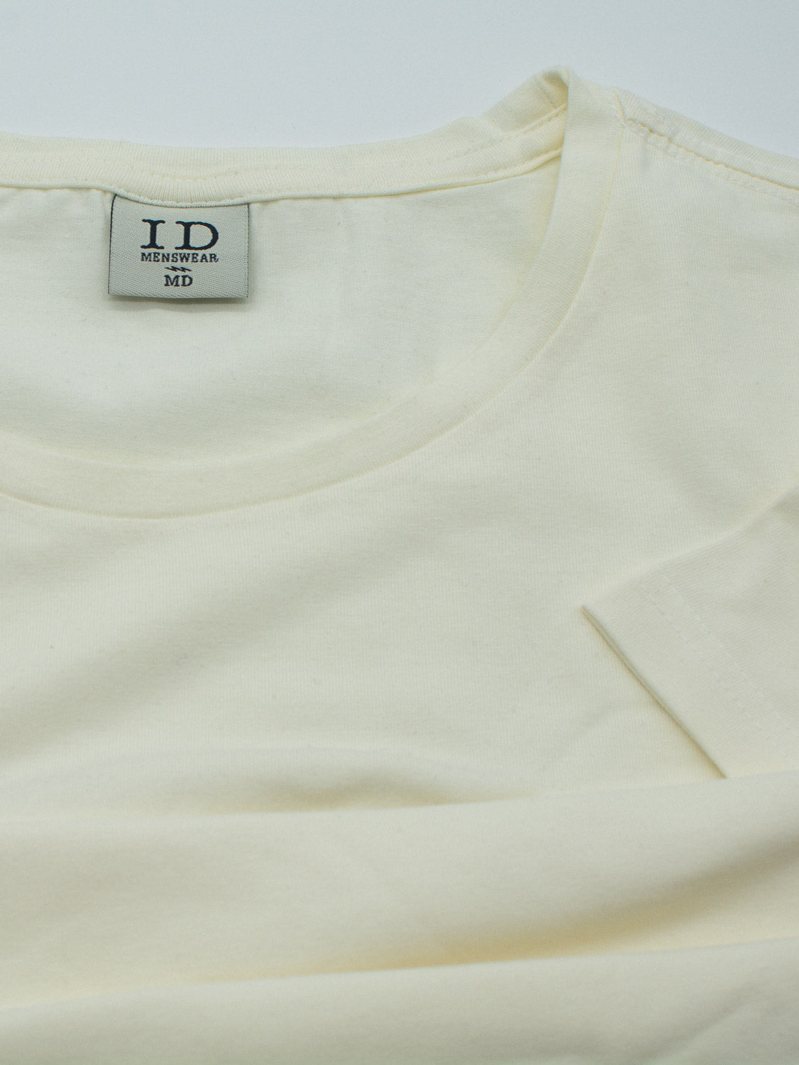 A cream-colored Form Crew Neck Tee from ID Menswear is shown spread flat, with the label visible inside the collar. The soft, slightly wrinkled fabric suggests a form-fitting design, emphasizing its comfort for everyday wear.