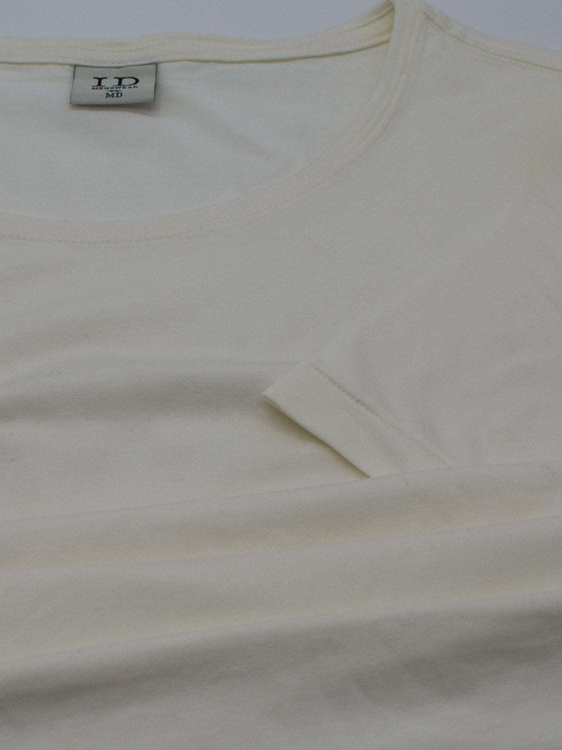 A close-up of a plain, light cream-colored T-shirt lying flat. This Slay Wide Neck Bamboo Blend Tee by ID Menswear is crafted from organic cotton and features a round neckline with visible fabric texture. A small label at the neck displays L in a dark font.