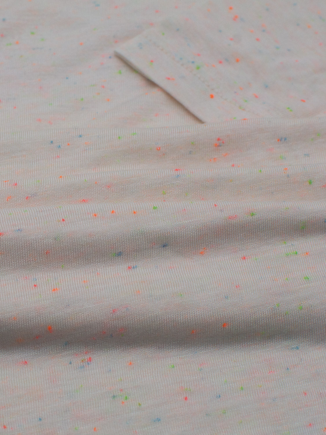 A light grey lightweight bamboo jersey from ID Menswears Outclass Bamboo Neppy Tee features a subtle ribbed texture with neon pink, green, blue, and yellow specks. The fabric is displayed as if to craft a crew neck t-shirt, with a small rectangular piece laid flat on top.