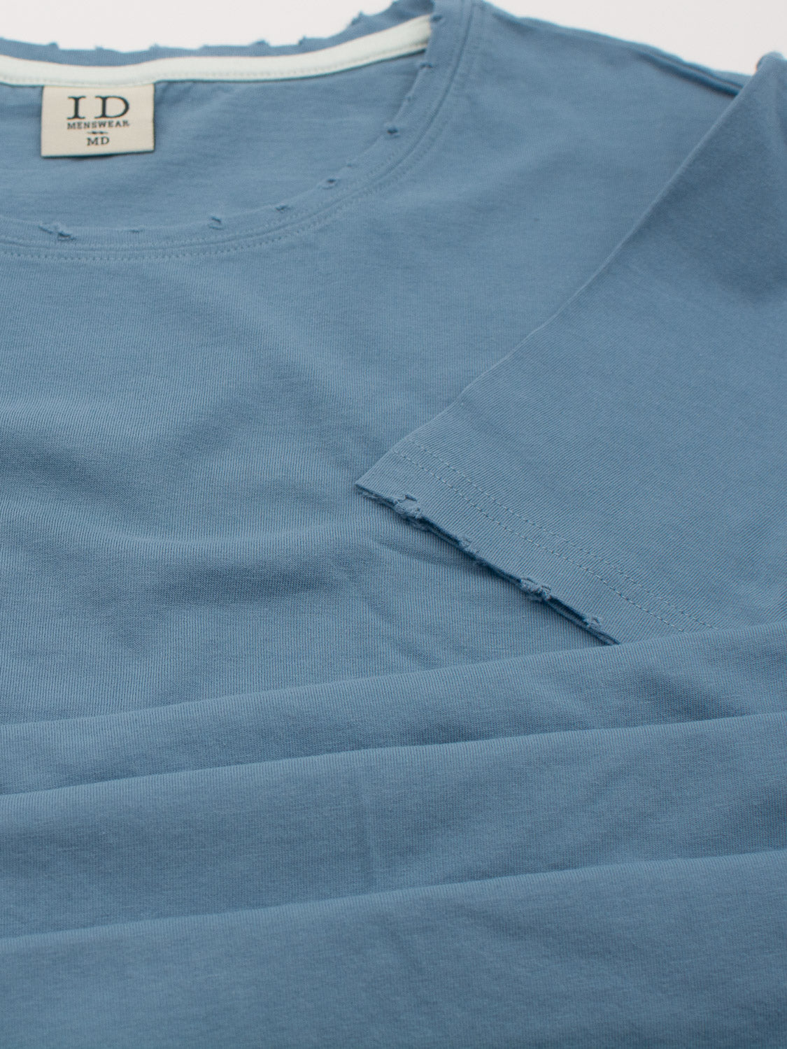 A blue Trekker Tattered Crew Neck Tee from ID Menswear, crafted from organic cotton with visible seam details, is folded neatly. The lightweight jersey fabric feels exceptionally soft, with a collar tag indicating MEDIUM.