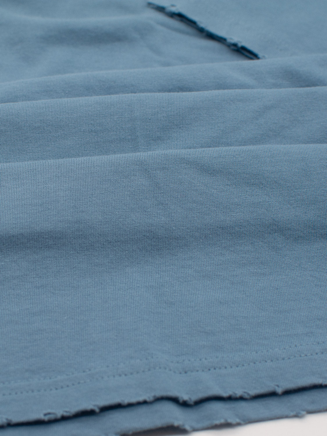 A close-up of folded blue fabric, showcasing its visible texture and weave, highlights the lightweight jersey of ID Menswears Trekker Tattered Crew Neck Tee, made from soft organic cotton with frayed edges for a gentle appearance.