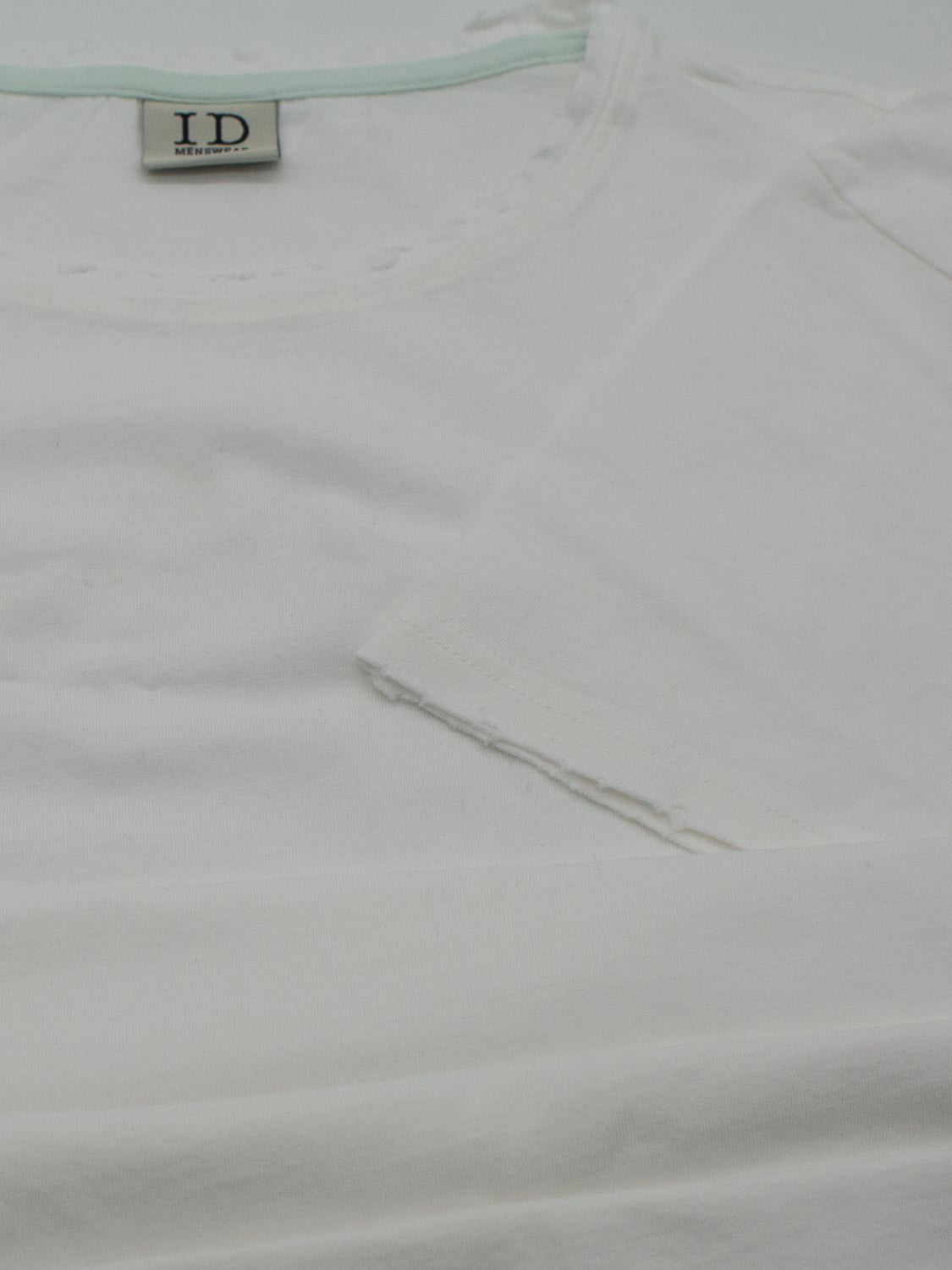 Close-up of a folded white Trekker Tattered Crew Neck Tee by ID Menswear. The label shows ID, see inside. Made from organic cotton, the jersey fabric appears soft and lightweight with a clean, simple design.