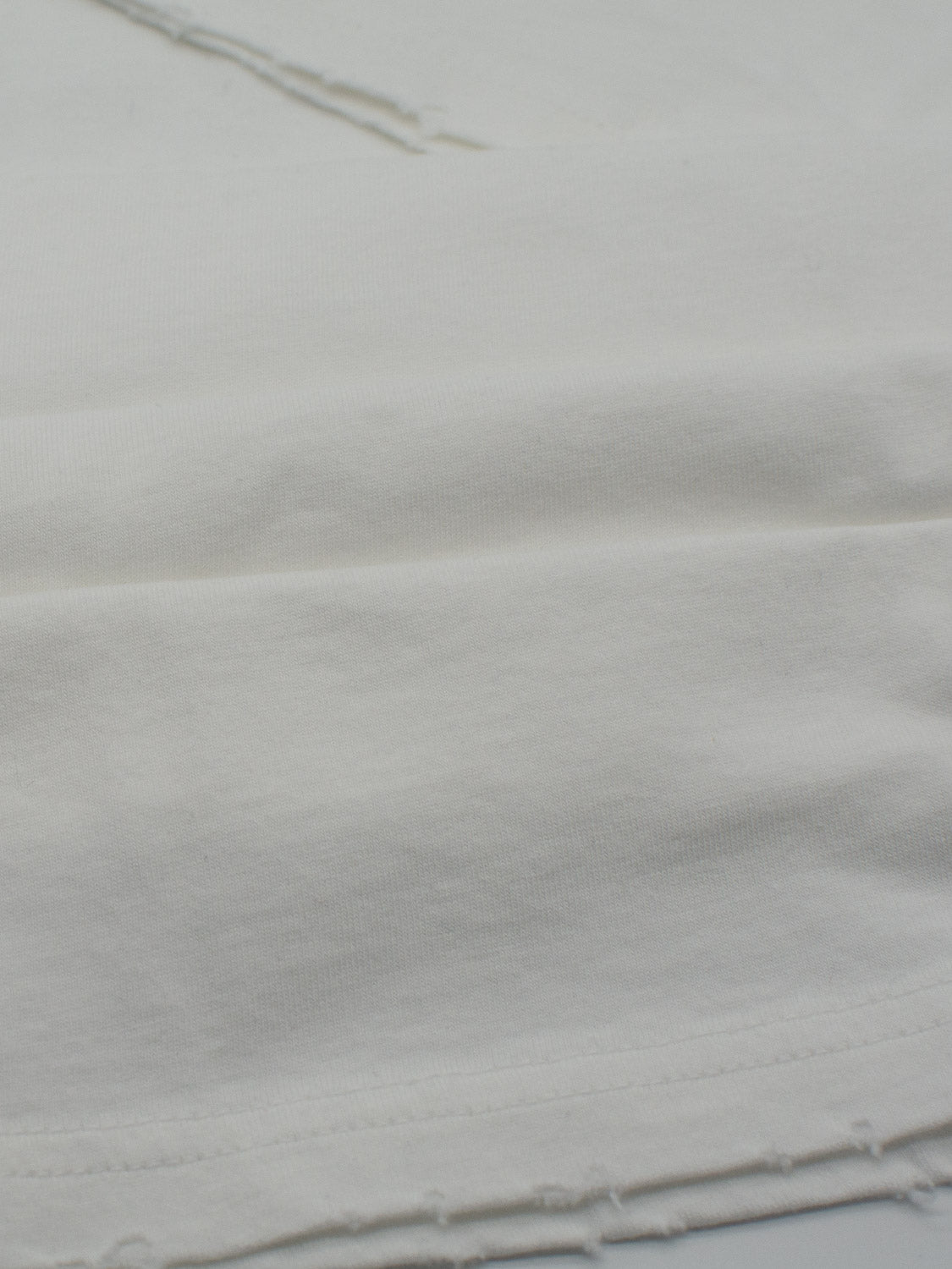 Close-up of an ID Menswear Trekker Tattered Crew Neck Tee in white, highlighting its textured surface from lightweight jersey. The slightly uneven edge and subtle diagonal lines showcase the softness of the organic cotton, with shadows emphasizing its texture and gentle folds.