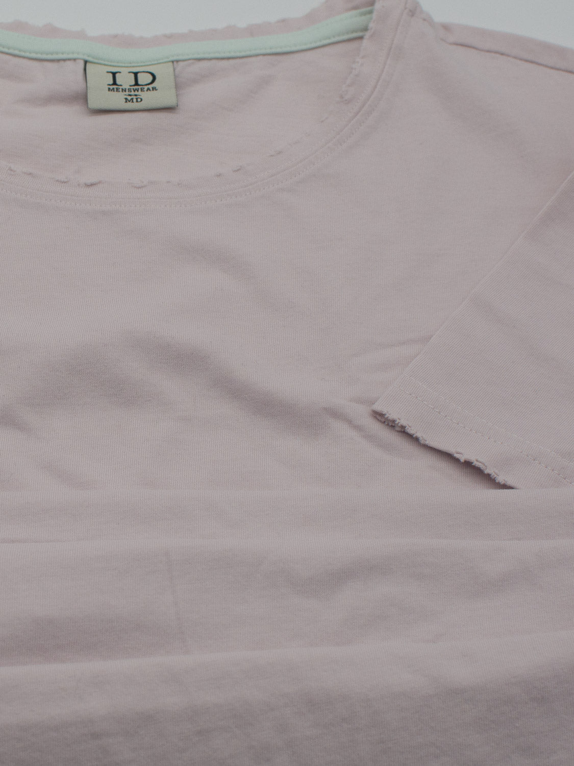 The Trekker Tattered Crew Neck Tee by ID Menswear is a light pink, lightweight jersey t-shirt made from organic cotton, featuring rough, unfinished edges on the collar and sleeves. It is tagged with the brands acronym and size inside.