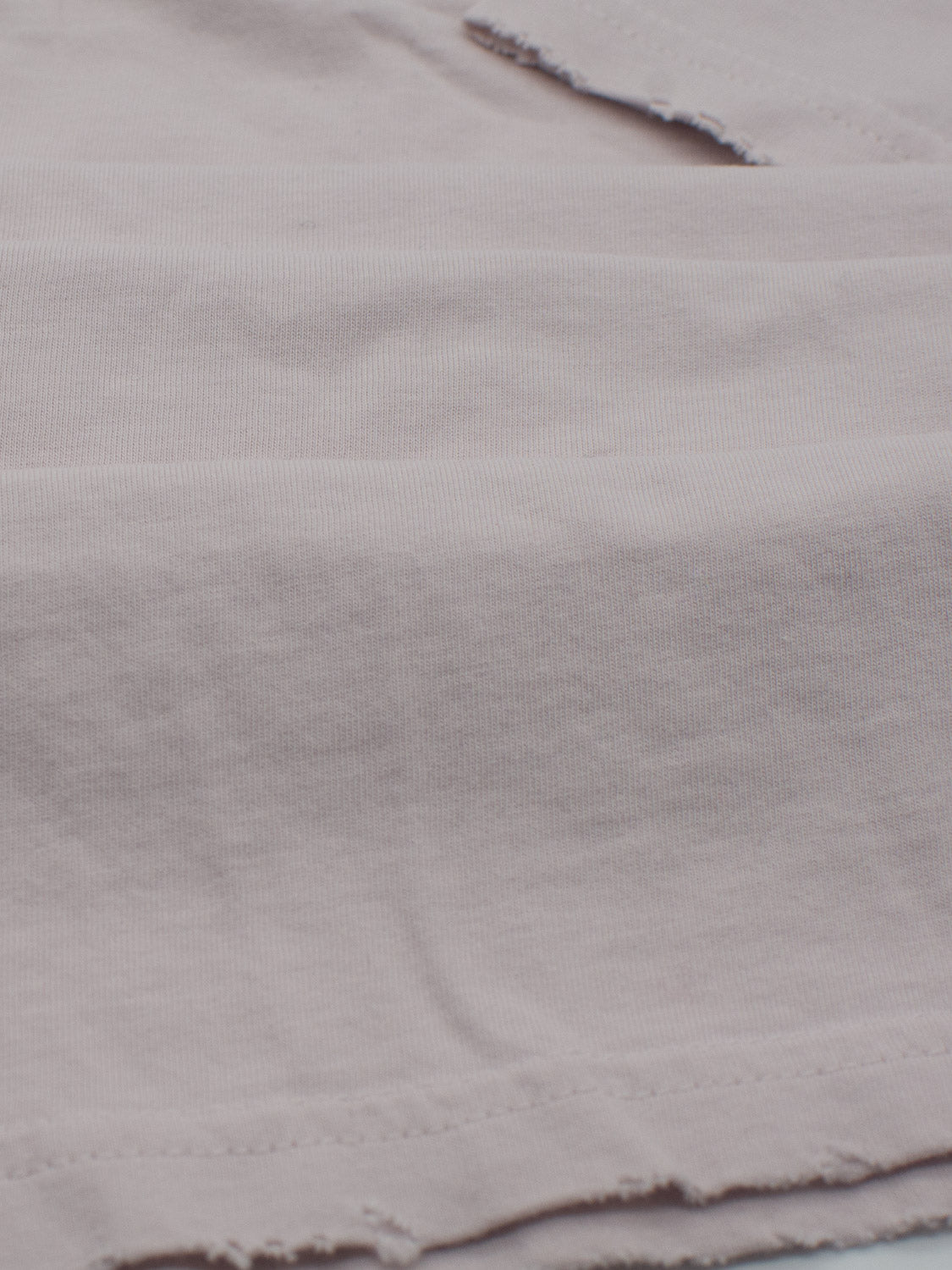 The Trekker Tattered Crew Neck Tee by ID Menswear features a close-up of light pink, frayed, organic cotton fabric. The visible texture and stitching, with slightly distressed edges, add a rustic touch to this lightweight jersey.