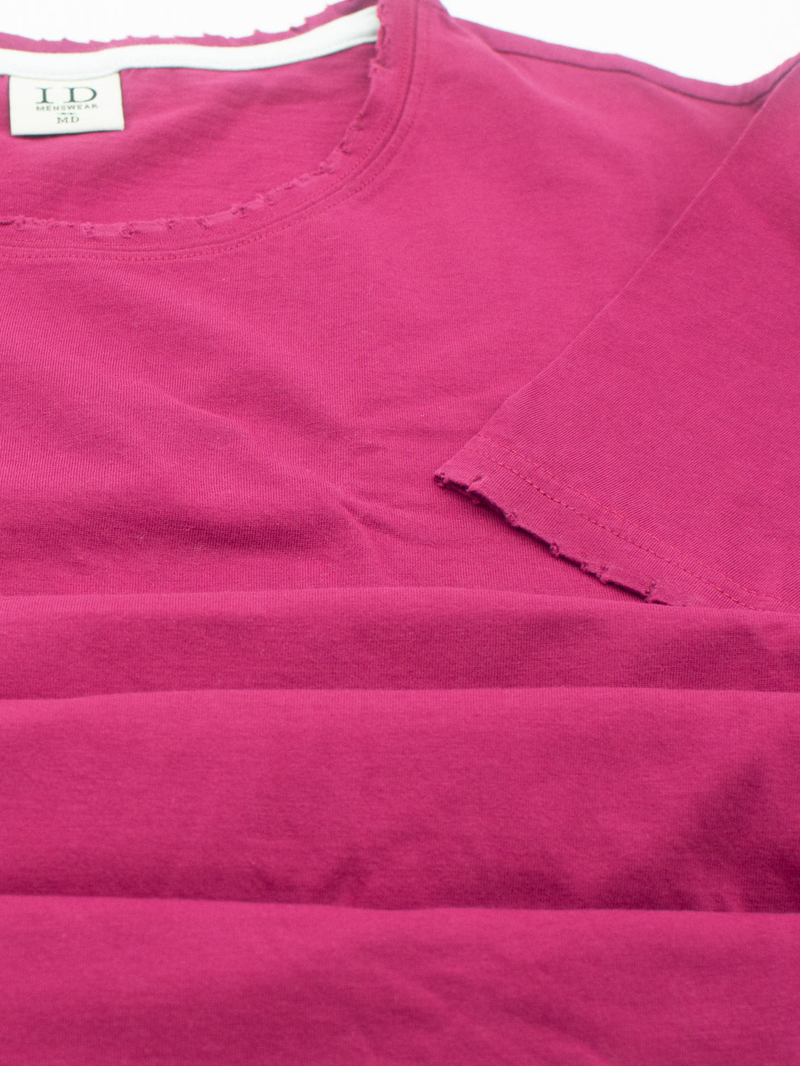 A close-up of a magenta Trekker Tattered Crew Neck Tee from ID Menswear is laid flat, showcasing its lightweight jersey fabric with a round neckline and short sleeves. A small label is visible at the neckline, and the smooth organic cotton fabric is brightly colored.