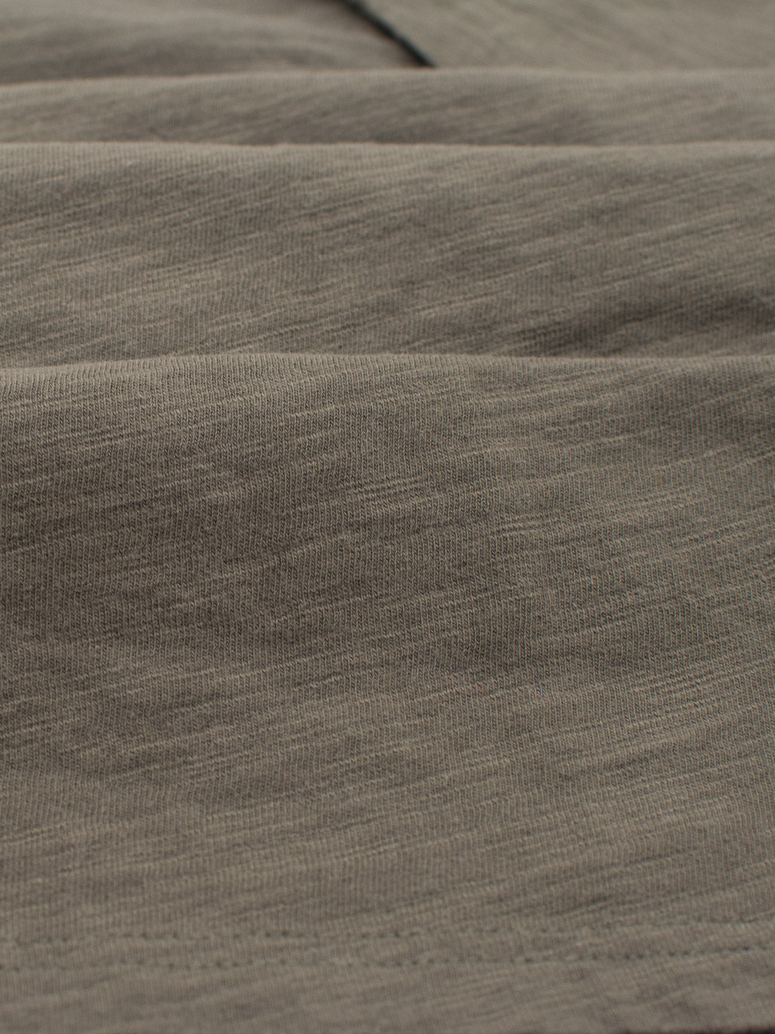 A close-up of the Mini Flip Slub Yarn Tee by ID Menswear showcases a textured olive green fabric similar to slub cotton, with visible stitching and subtle folds. This soft, slightly wrinkled material exudes an effortless casual look, enhanced by light and shadow playing on its intricate texture.