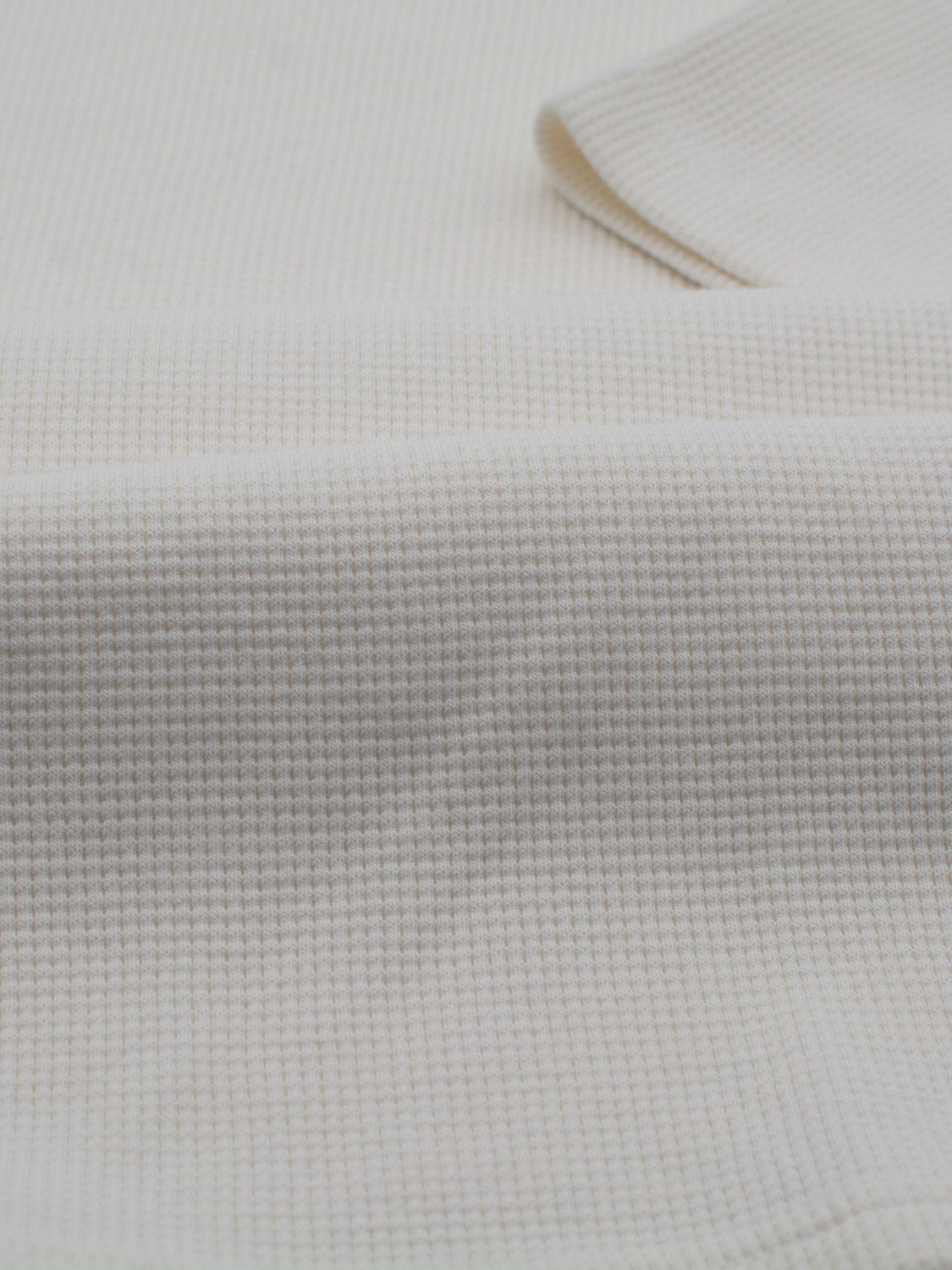 Close-up of the off-white, grid-patterned fabric of the Urban Waffle Knit Tee by ID Menswear, made from a soft cotton-spandex blend. The textured material appears slightly gathered, forming gentle waves and shadows ideal for relaxed casual wear.