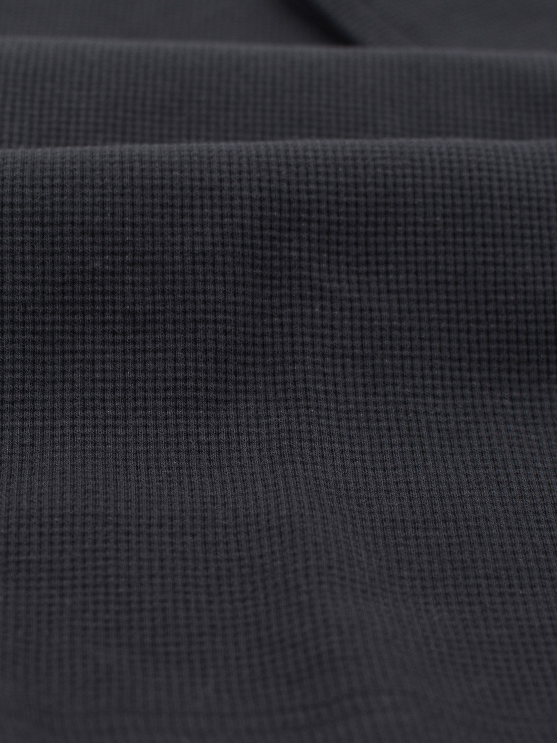 A close-up of ID Menswears Urban Waffle Knit Tee reveals its dark gray, textured fabric with a grid-like waffle pattern. The cotton-spandex blend gives it a soft and slightly undulating surface, suggesting subtle folds or waves.