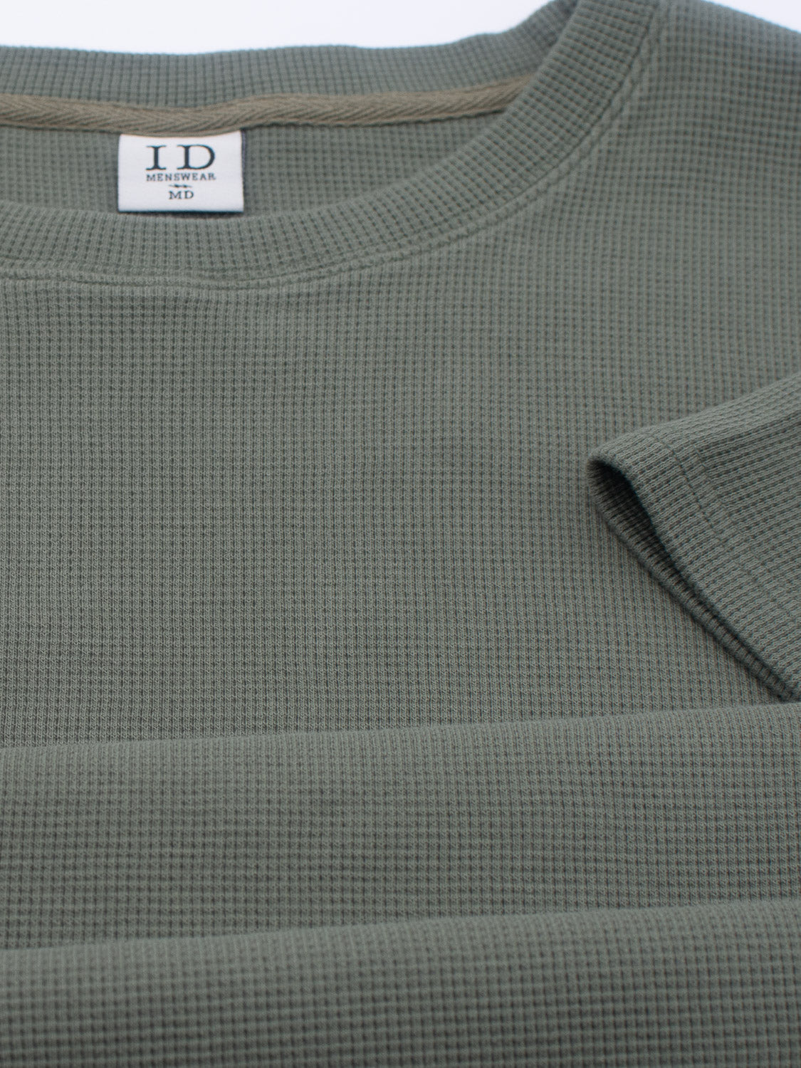 Close-up of the Urban Waffle Knit Tee: a ribbed, olive-green waffle knit with a round neckline. A label inside reads ID Menswear M. Perfect for casual wear, it’s crafted from a cozy cotton-spandex blend that enhances its textured design.
