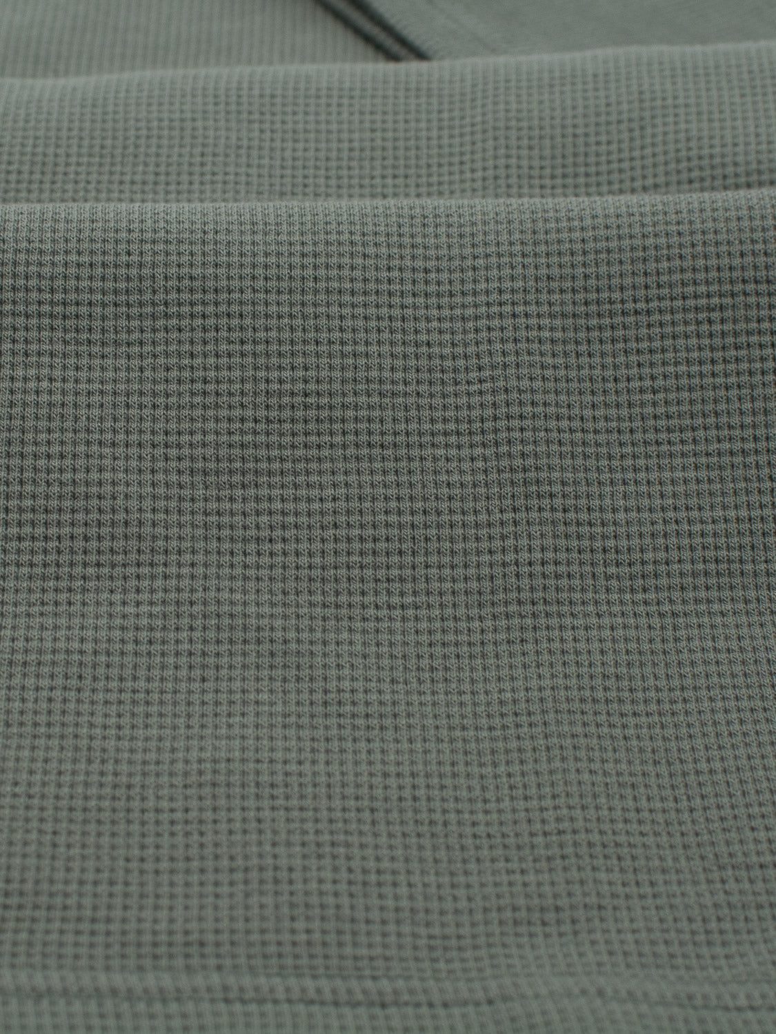 A close-up shot of the green Urban Waffle Knit Tee by ID Menswear highlights its subtle waffle-knit grid pattern. The cotton-spandex blend feels soft and textured, with gentle folds casting shadows for a muted, comforting tone.