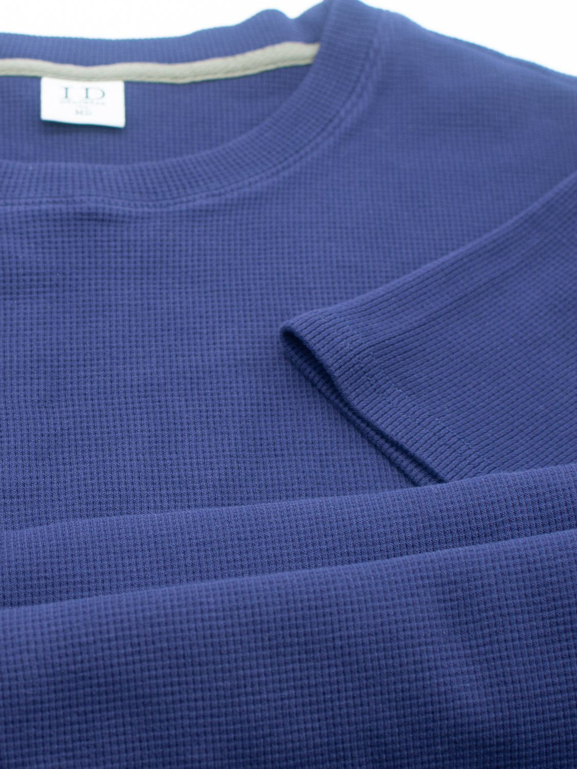 A close-up of the Urban Waffle Knit Tee in blue, featuring a folded design with a waffle knit pattern. The neckline label displays ID, indicating its a medium-sized piece made by ID Menswear from a soft cotton-spandex blend, promising comfort and effortless casual wear.