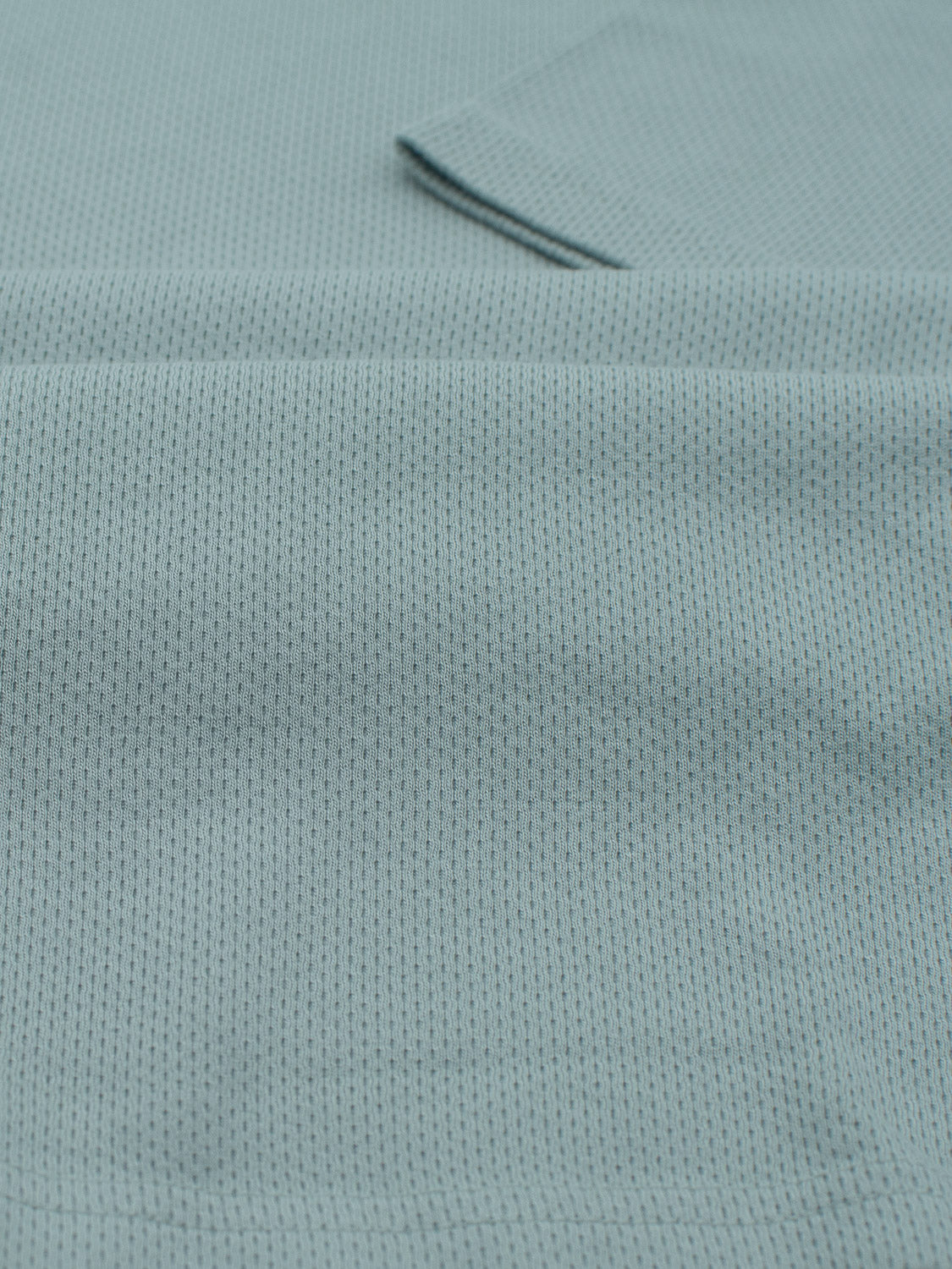 A close-up of ID Menswears Urban Honeycomb Mesh Tee displays a textured light blue fabric with dark speckles. The soft and breathable material, hinting at a bamboo-organic cotton blend, features subtle folds and creases for flexibility and comfort.