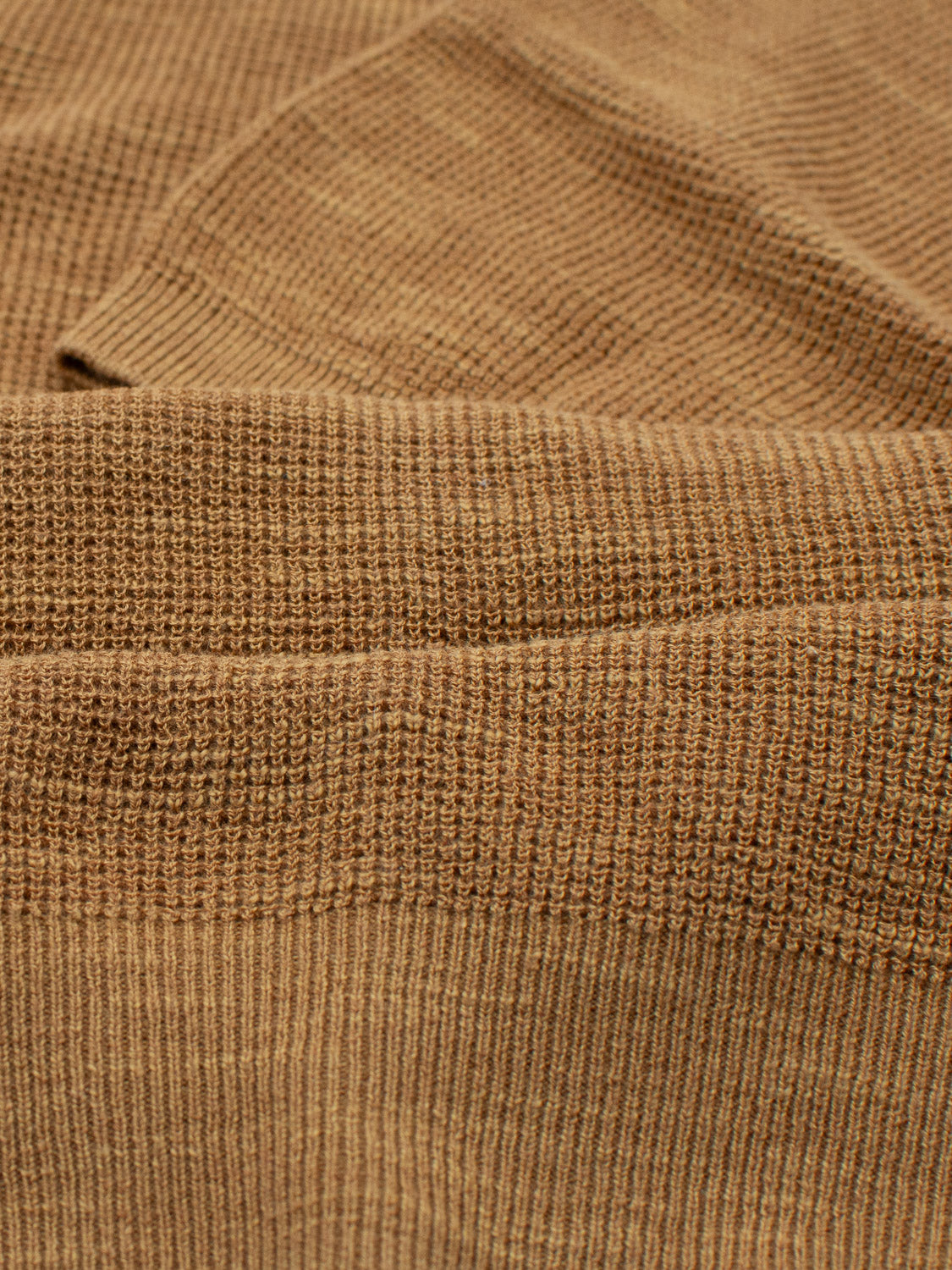 Close-up of textured brown fabric from ID Menswears Ankara Linen Blend Waffle Knit Polo, showcasing ribbed pattern with subtle color variations. The folds create shadows and highlights, emphasizing the lightweight, breathable texture ideal for warm weather.