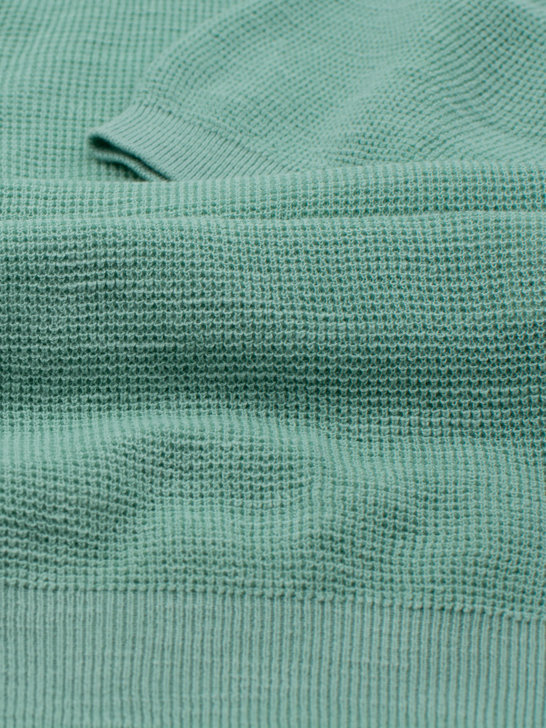 A close-up of a teal knitted fabric with a waffle texture resembles the Ankara Linen Blend Waffle Knit Polo by ID Menswear. The gently folded material highlights its thick, soft look while being lightweight and breathable for warm weather.