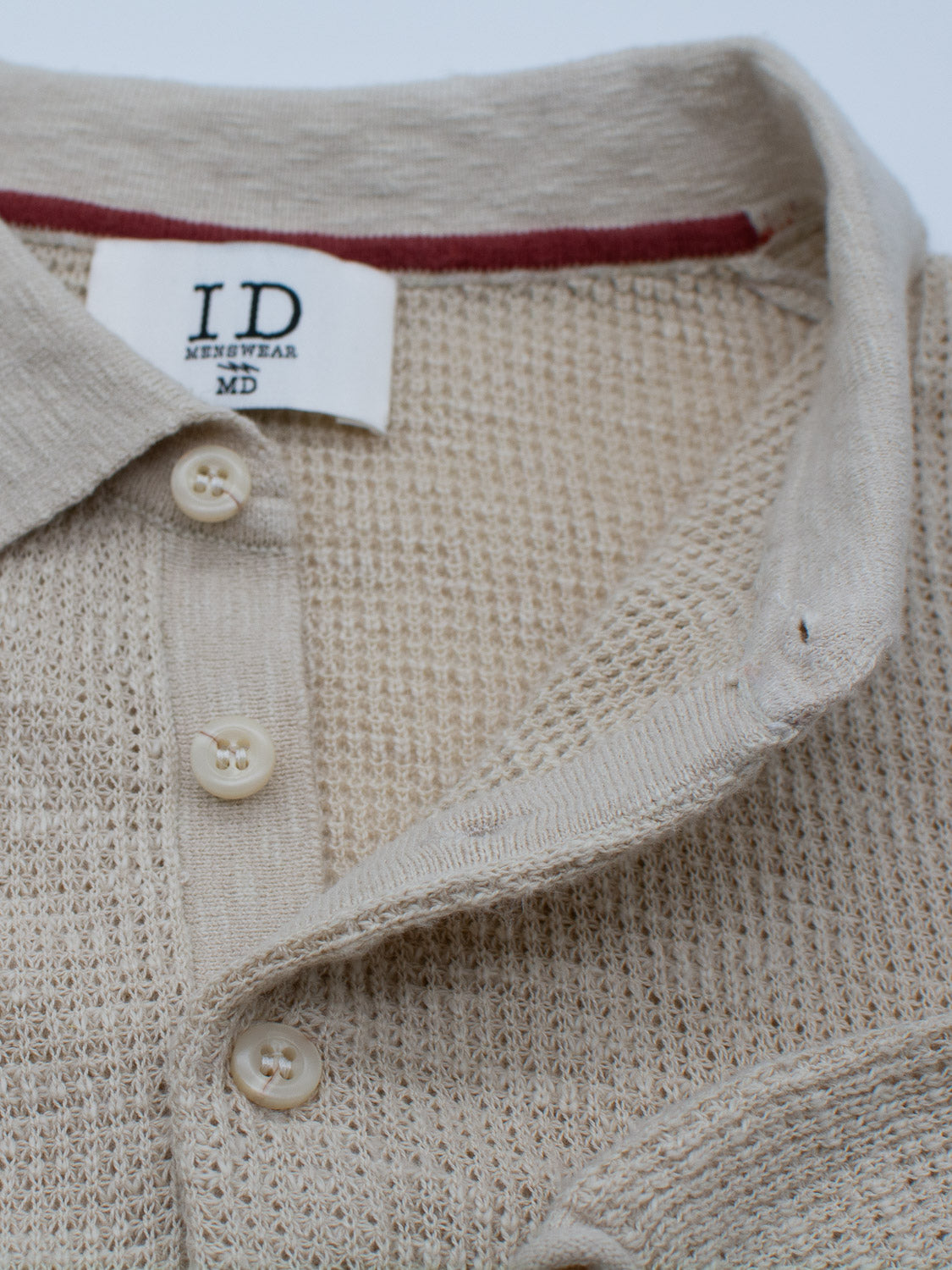 Close-up of the Athens Waffle Knit Polo by ID Menswear, highlighting its beige waffle texture with a collar and three visible beige buttons. Inside, a white tag reads ID Menswear and MD, with a red fabric line adding to its casual style.