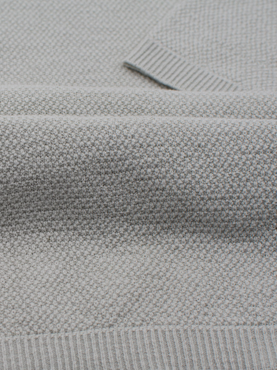 Close-up of a folded Austin Knitted Cotton Polo by ID Menswear in a textured light gray knit with visible ribbed cuffs. The dense, small-scale weave offers subtle uniformity and ensures breathable comfort, ideal for casual wear.