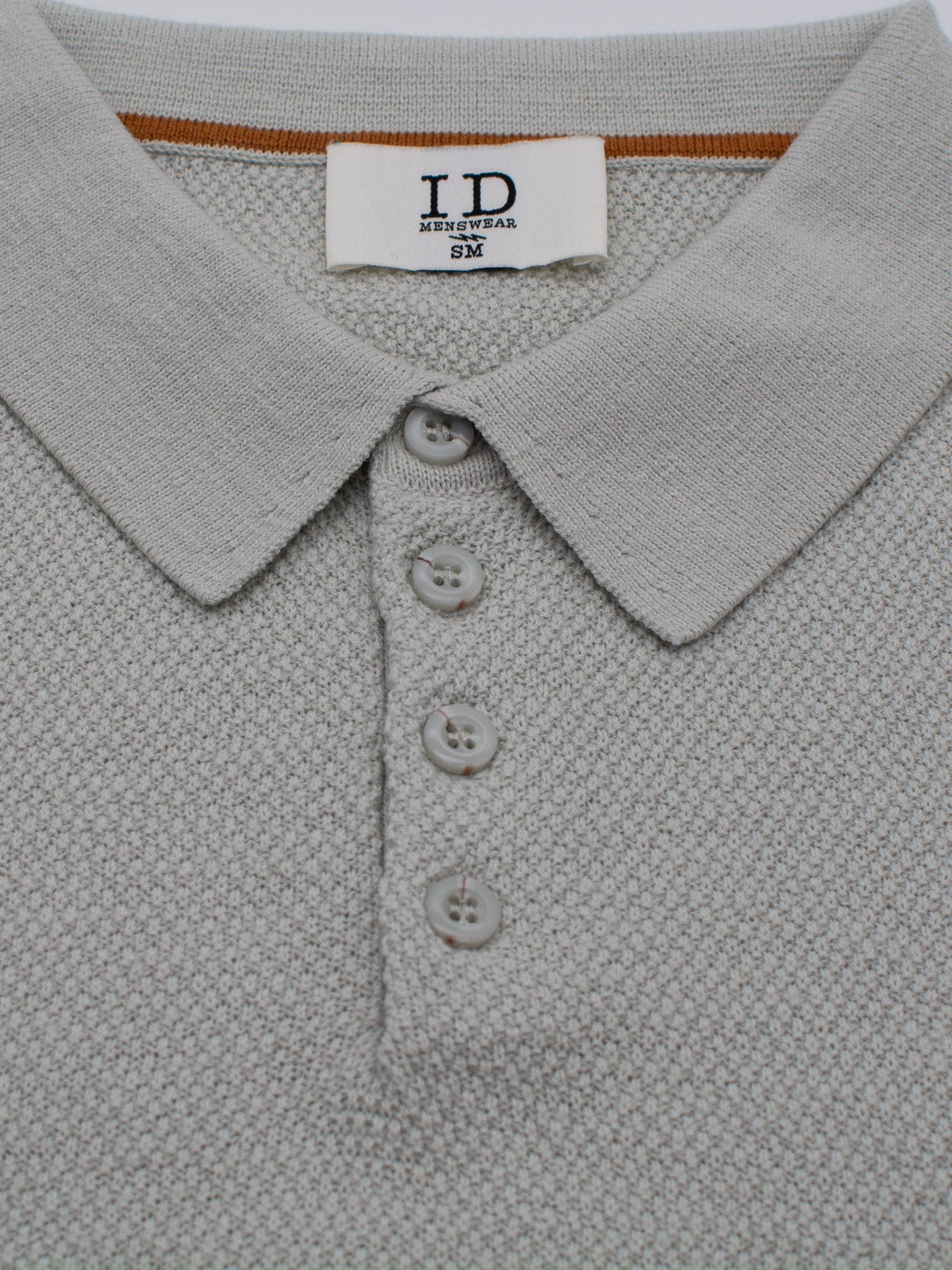 The close-up showcases the Austin Knitted Cotton Polo, a gray textured shirt by ID Menswear in size 3M. It features a collar with four buttons, an orange accent stripe, and a breathable fit for casual elegance and comfort.